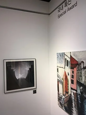 Two paintings hanging on a white gallery wall, with text in English and Korean indicating a special award.exhibited in Seoul Korea.