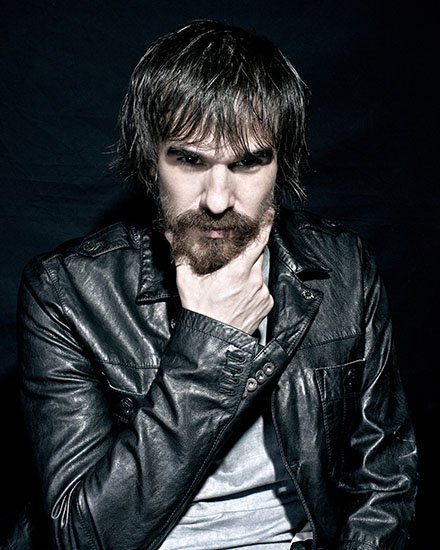 A man with dark hair and a beard wearing a black leather jacket, posing with his hand on his chin against a dark background.PHE. Bienal Fundacion Once