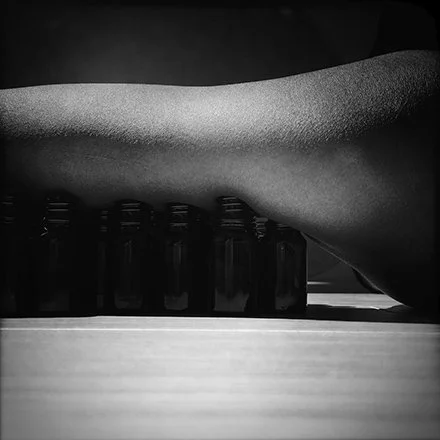 Close-up of a human arm resting on a row of small bottles on a flat surface, black and white photo.