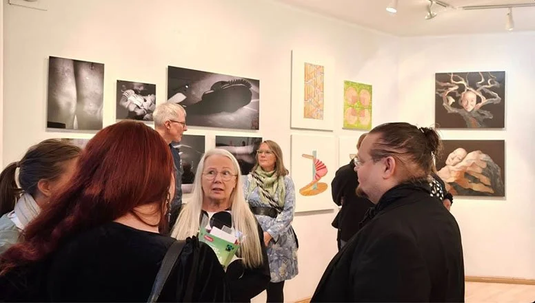 Group of people, including older women and men, in an art gallery discussing various black-and-white and colorful artwork displayed on the white walls.Life Sentence opening exhibition in Helsinki, Finland.