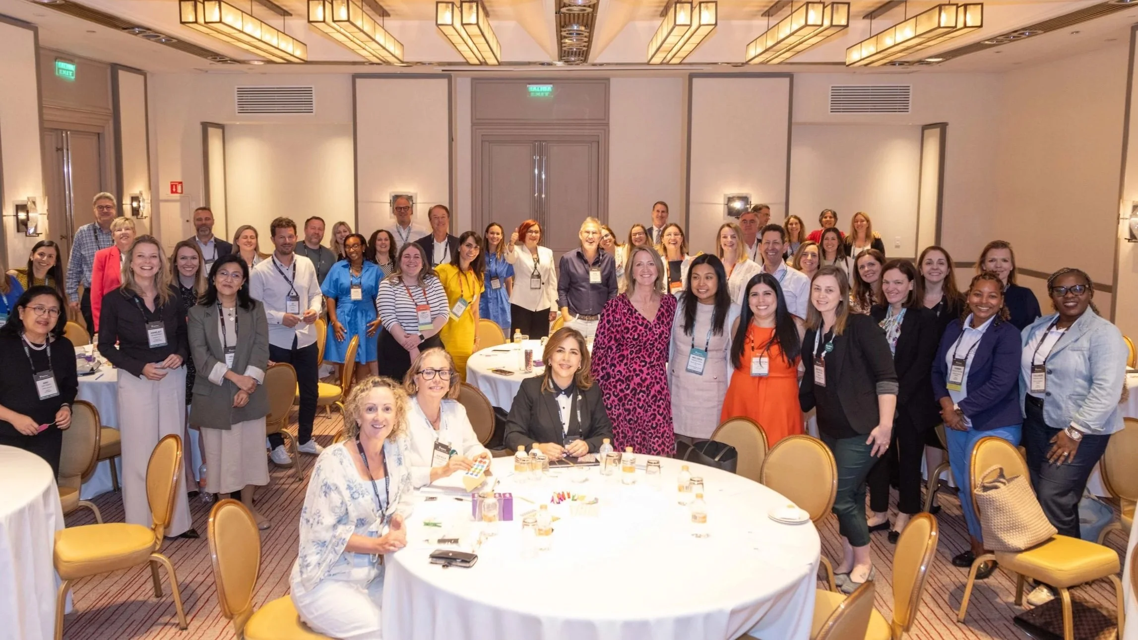Group of diverse people gathered around banquet tables at a conference, smiling for a photo in a banquet hall.