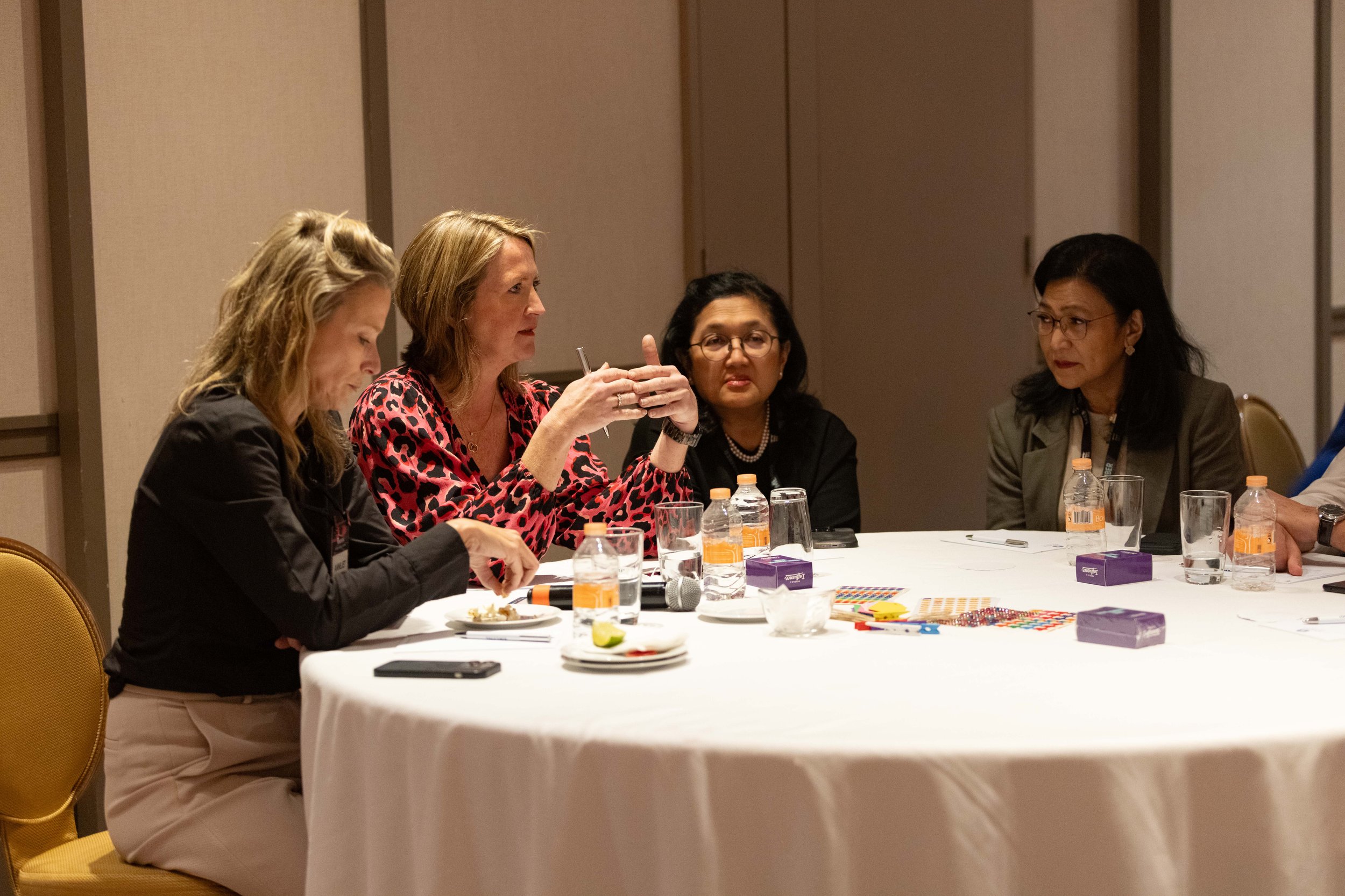 Group of women sitting at a round table during a meeting or conference, with beverages, snacks, and cards on the table.