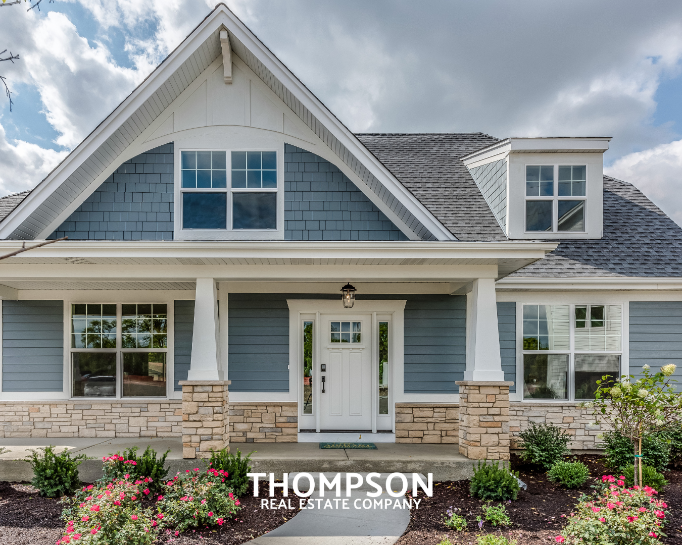 Front view of a modern house with blue siding, large windows, and a covered porch with a white door. The house has a gabled roof with gray shingles, and there are flowerbeds with pink and green plants in the foreground. The image has a logo at the bottom that says "Thompson Real Estate Company."
