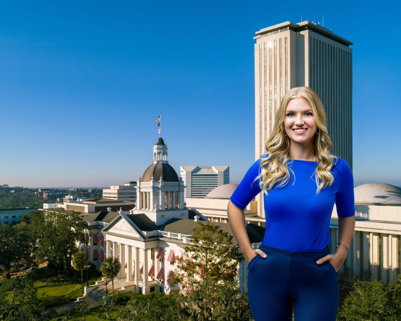 A woman with blonde hair smiling, wearing a blue top and dark blue pants, standing outdoors with a cityscape background featuring a tall modern building and a historic building with a dome and flags.