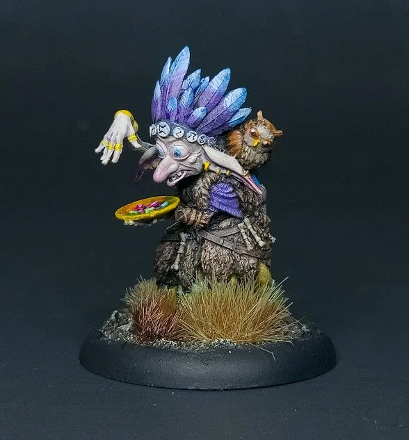 Miniature figure of a wizard with a staff, large feathered headdress, and a feline companion, standing on terrain with grass.