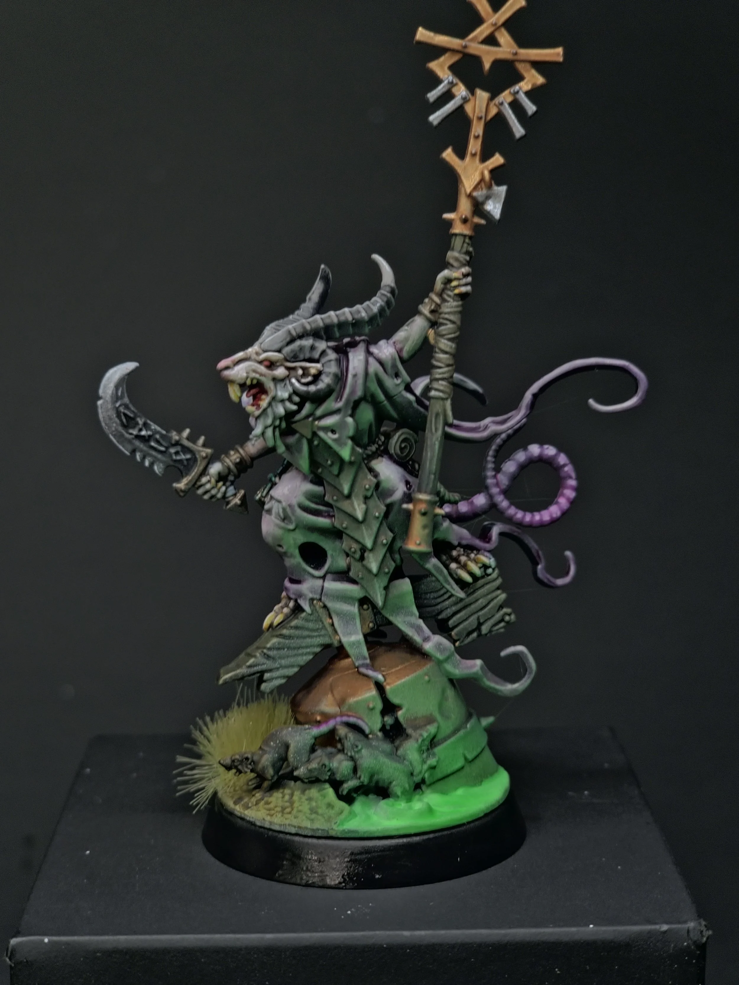A detailed miniature figurine of a fantastical creature with a dragon-like head, horns, and a tail, wielding a spear and wearing armor, standing on a small terrain base with grass and a creature at its feet, painted in various shades of green, gray, 