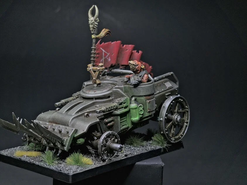 Miniature model of a futuristic armored vehicle with spikes, guns, with a rat figure inside and a red flag with a crack across like a break, on a display base with grass accents.