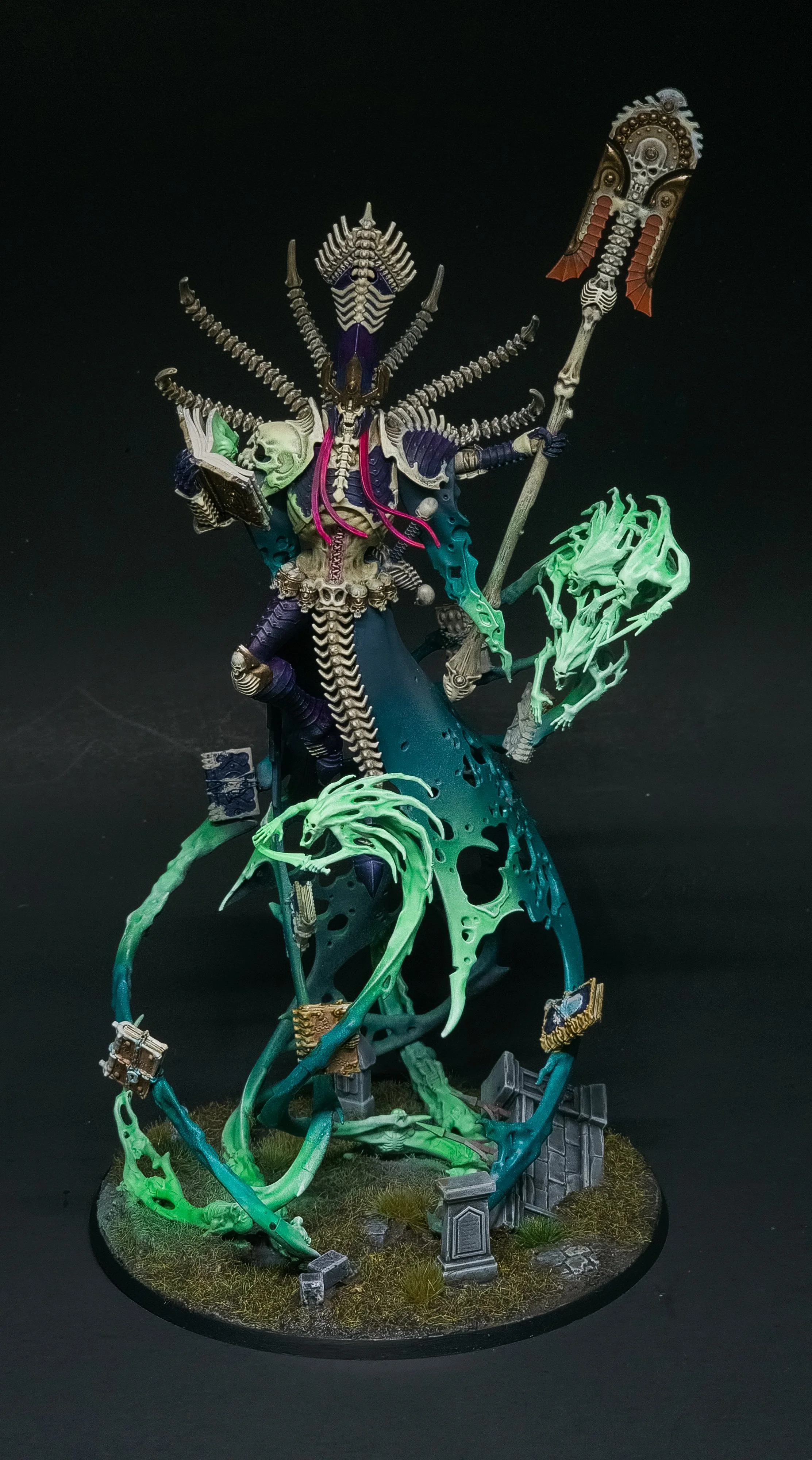 A detailed miniature figurine of a fantasy undead sorcerer or necromancer with a skeletal appearance, multiple skulls, and tentacle-like structures, holding a staff with a skull and wing detail, standing on a textured base with grass and broken stone elements.