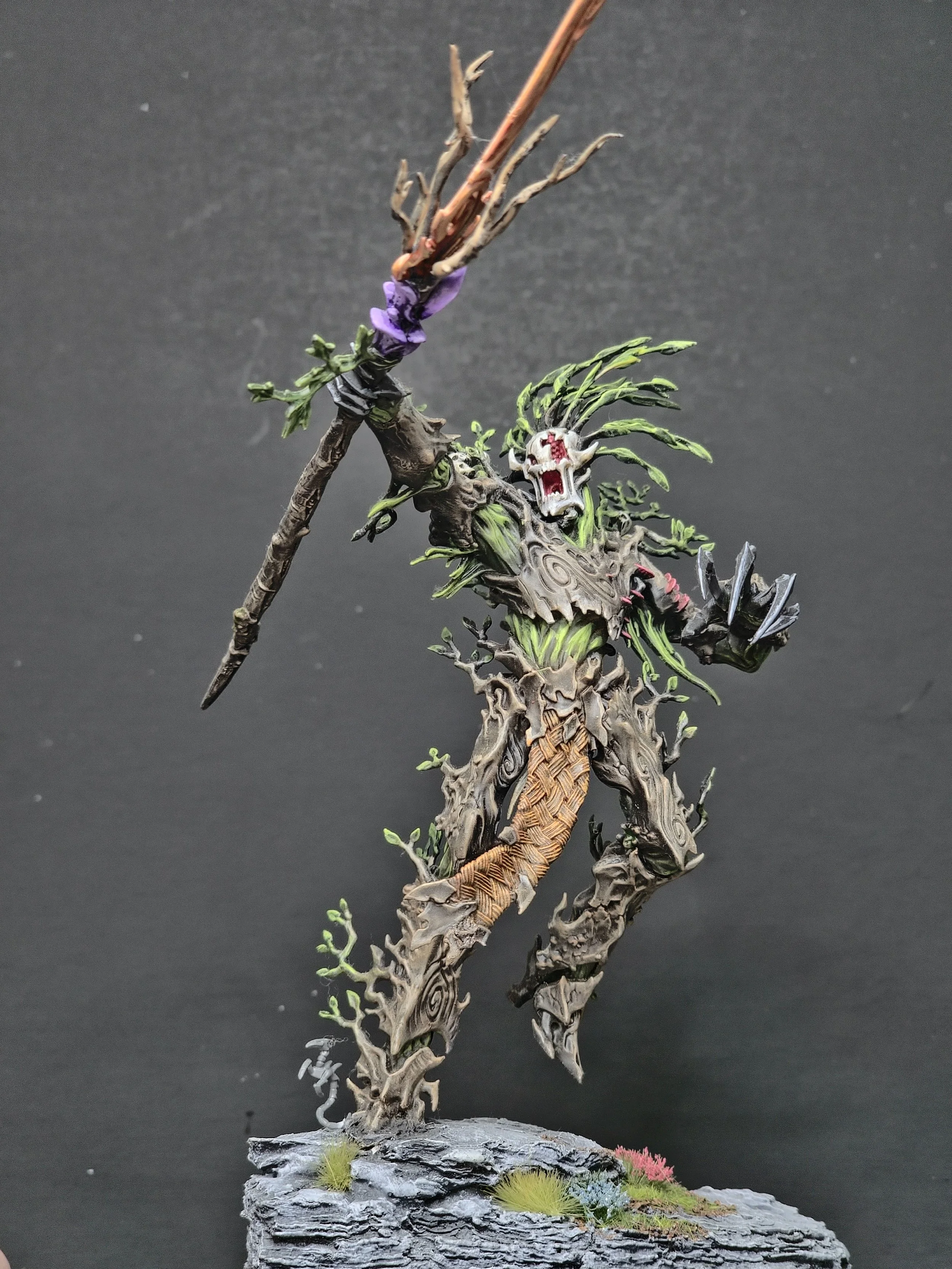 A detailed miniature sculpture of a mythical creature with a dragon-like body, adorned with green foliage and a skull on its head, standing on a textured rocky base with small tufts of grass.