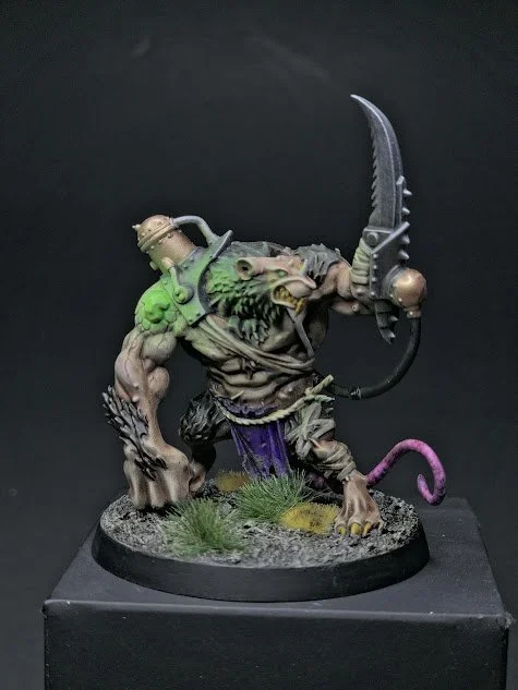 A fantasy monster miniature figurine with a muscular humanoid body, a lion-like head, and a tail, holding a curved blade. It features metallic details, green shoulder armor, and purple cloth, mounted on a textured base with grass.