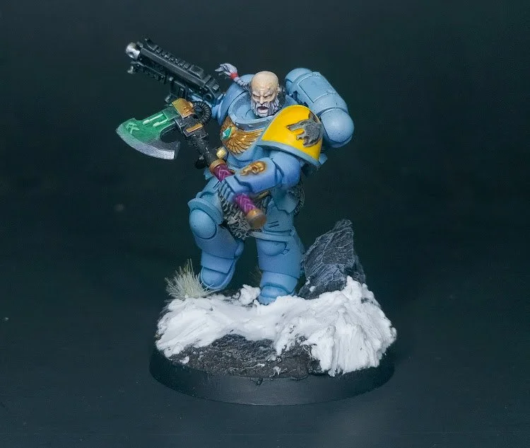A detailed painted miniature figure of a Space Marine from Warhammer 40,000, posed on a snow-covered rocky base, holding a large gun and a sword.