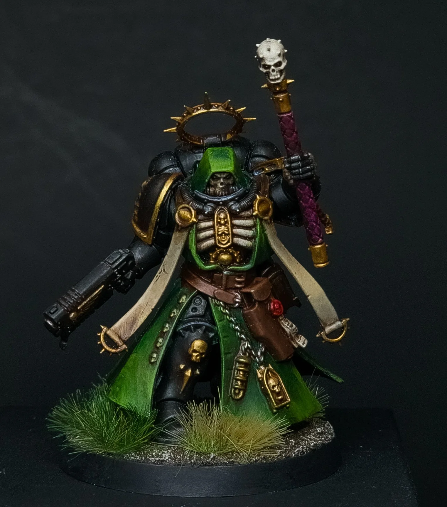 A detailed miniature figurine of a fantasy warrior wearing black and green armor, wielding a staff with a skull on top, standing on a base with grass and rocks.