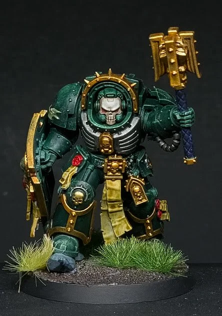 Miniature figurine of a green armored warrior with gold details, holding a large hammer, standing on a grassy base.