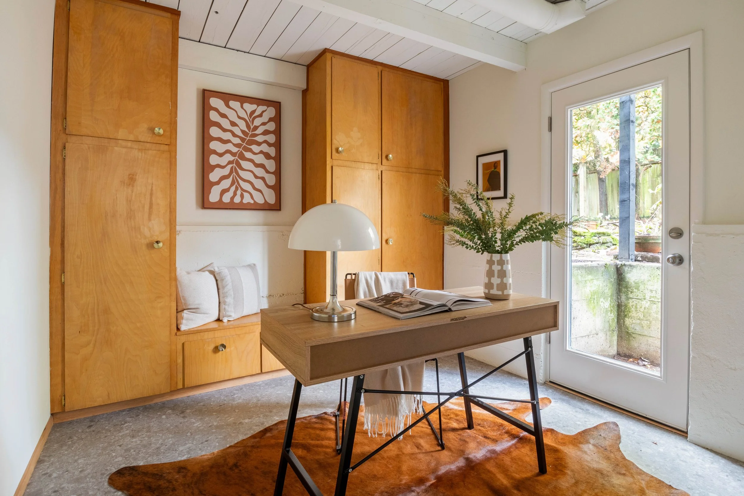 Interior view of a room showing wooden cabinets, a window door with outdoor greenery, a table with a plant in a patterned vase, a white table lamp, and a book, with an orange rug on the floor.