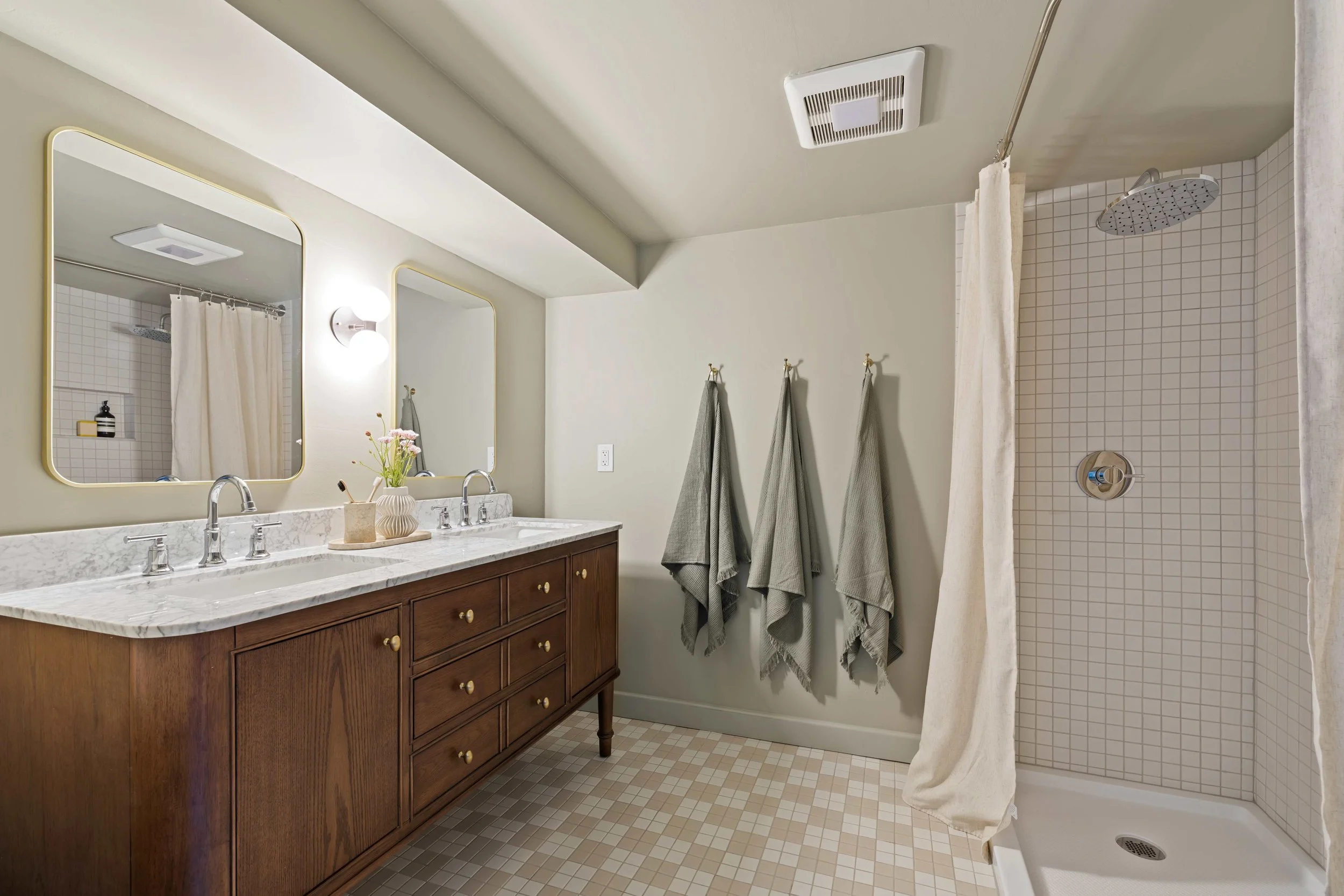 Bathroom with double sink vanity, large mirror, towels hanging on wall, shower with curtain, ceiling vent, and tile flooring.