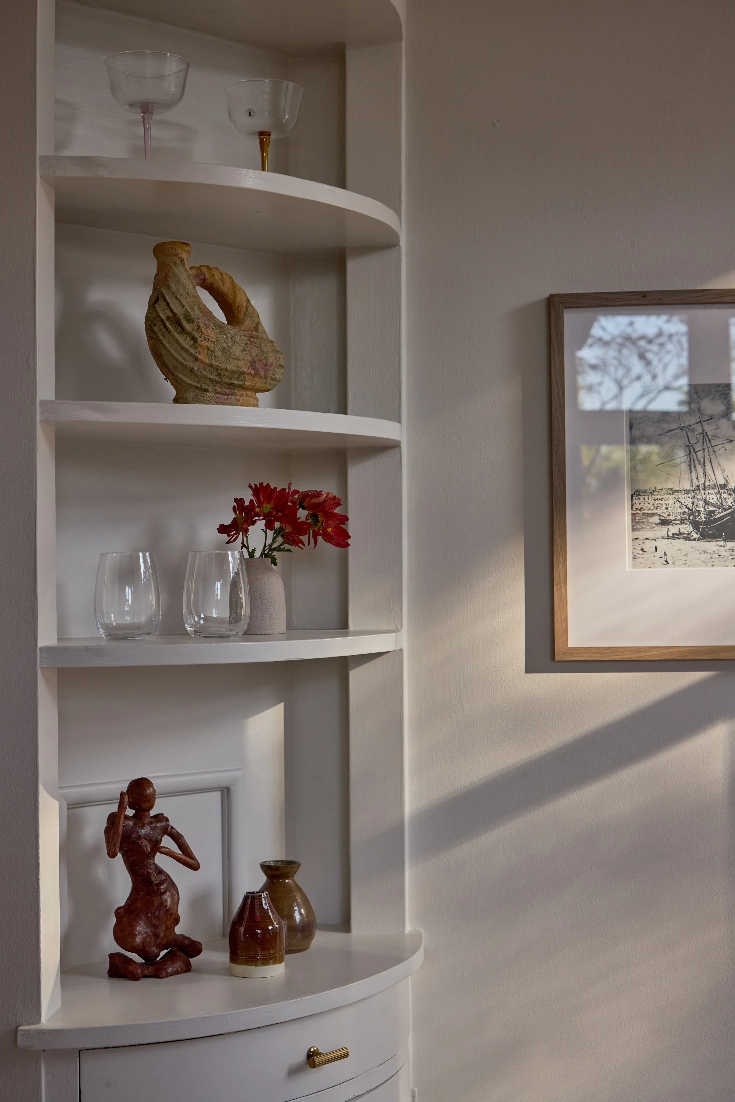 A white display cabinet with three shelves holding various decorative objects: glassware on the top, a ceramic sculpture resembling a bird or seashell on the middle, and vases and a red flower arrangement on the lower shelf. A framed artwork is partially visible on the wall beside the cabinet, and shadows from sunlight are cast across the wall.