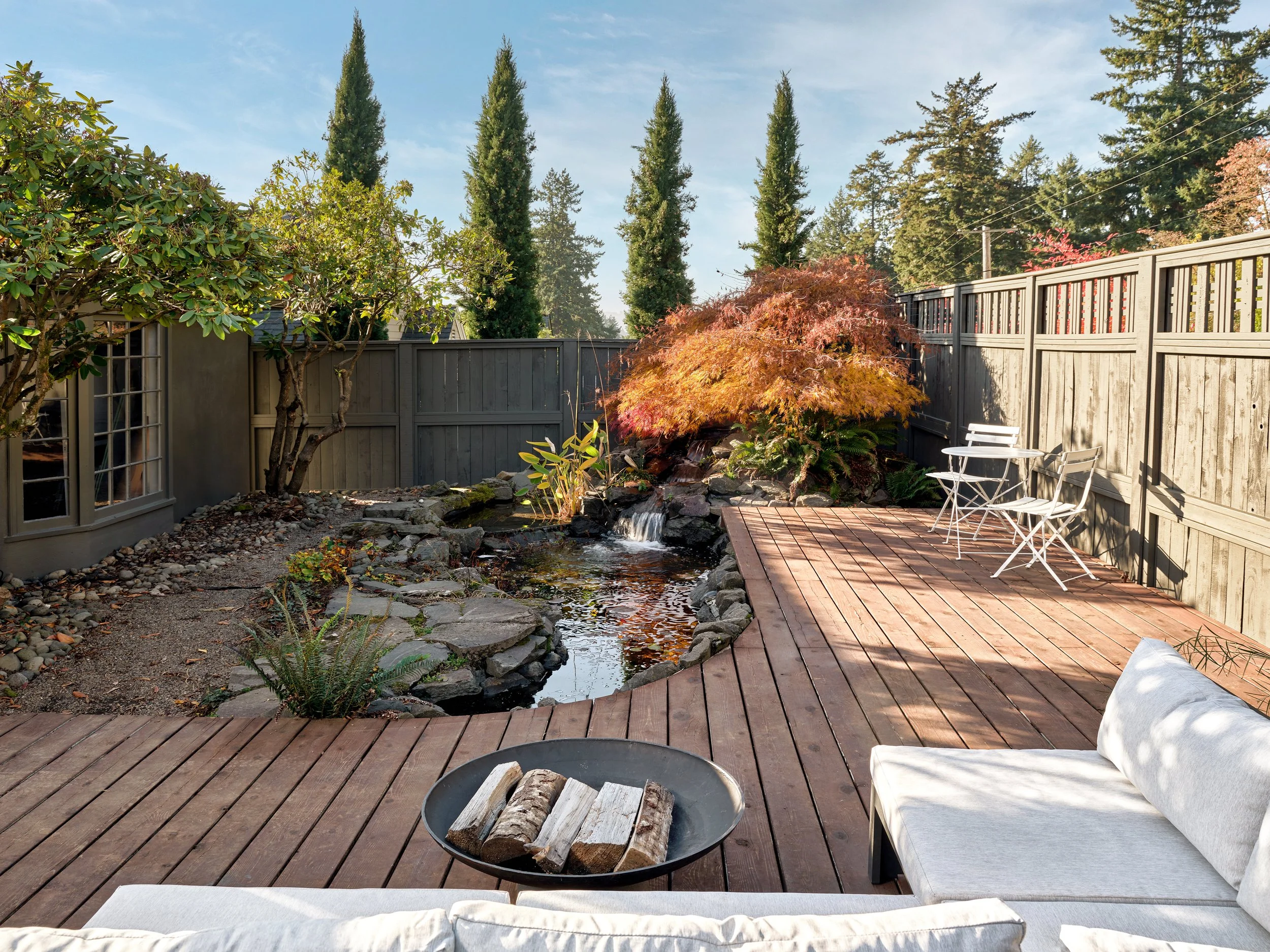 Backyard garden with wooden deck, outdoor seating, a small waterfall pond, trees, and a gray fence.