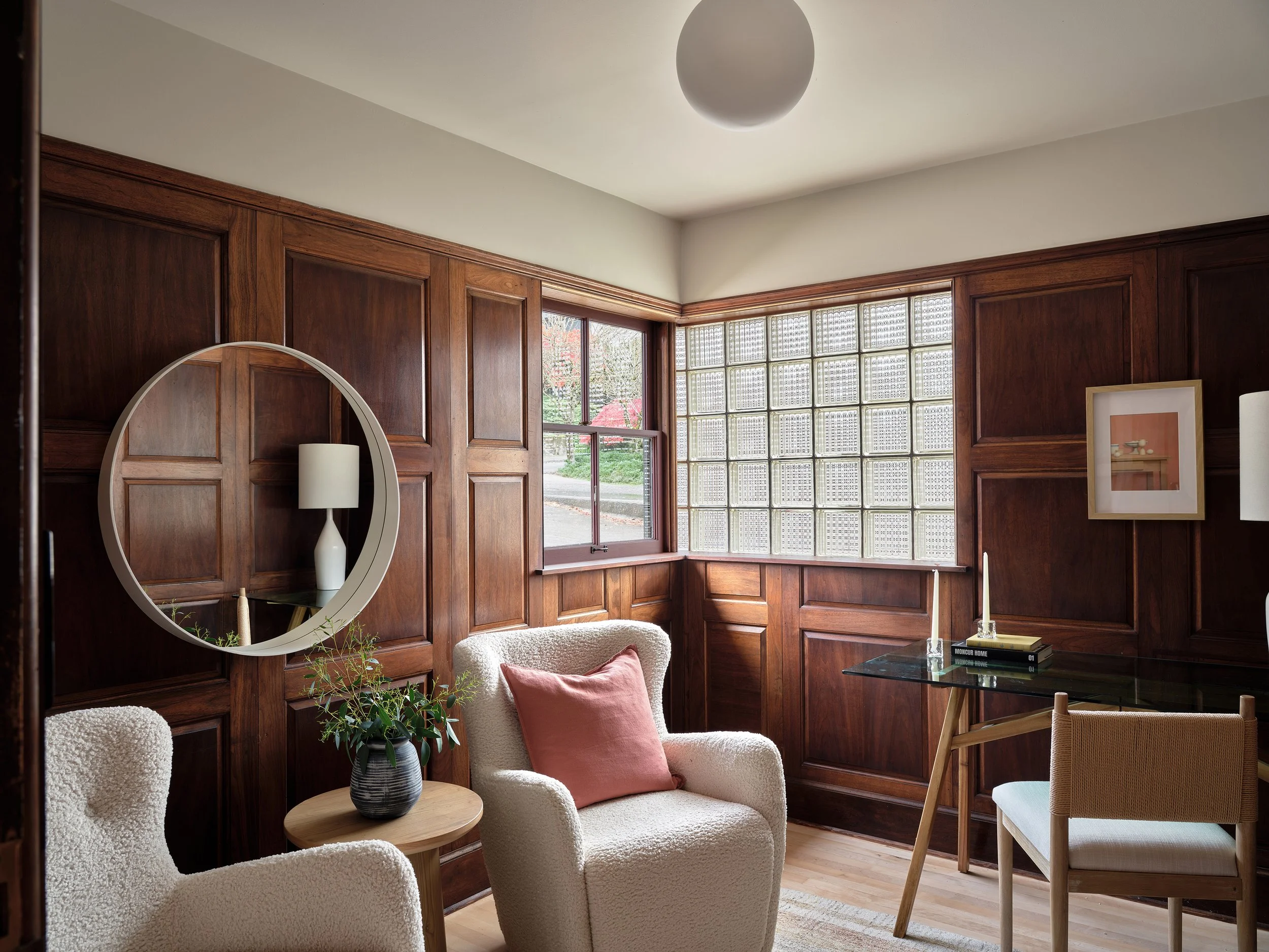 Cozy interior with wood-paneled walls, a window with glass blocks, and furniture including white armchairs, a side table with a plant, a mirror, and a bookshelf.