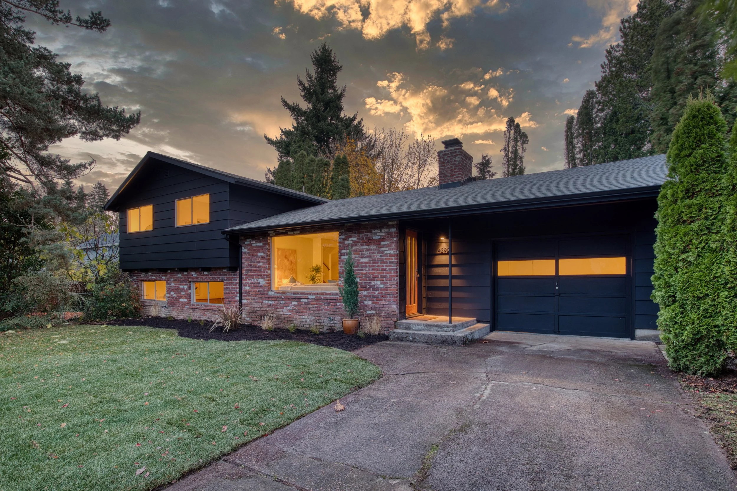 A house at dusk with a brick and black exterior, illuminated windows, and a garage door, surrounded by trees and a grassy yard.