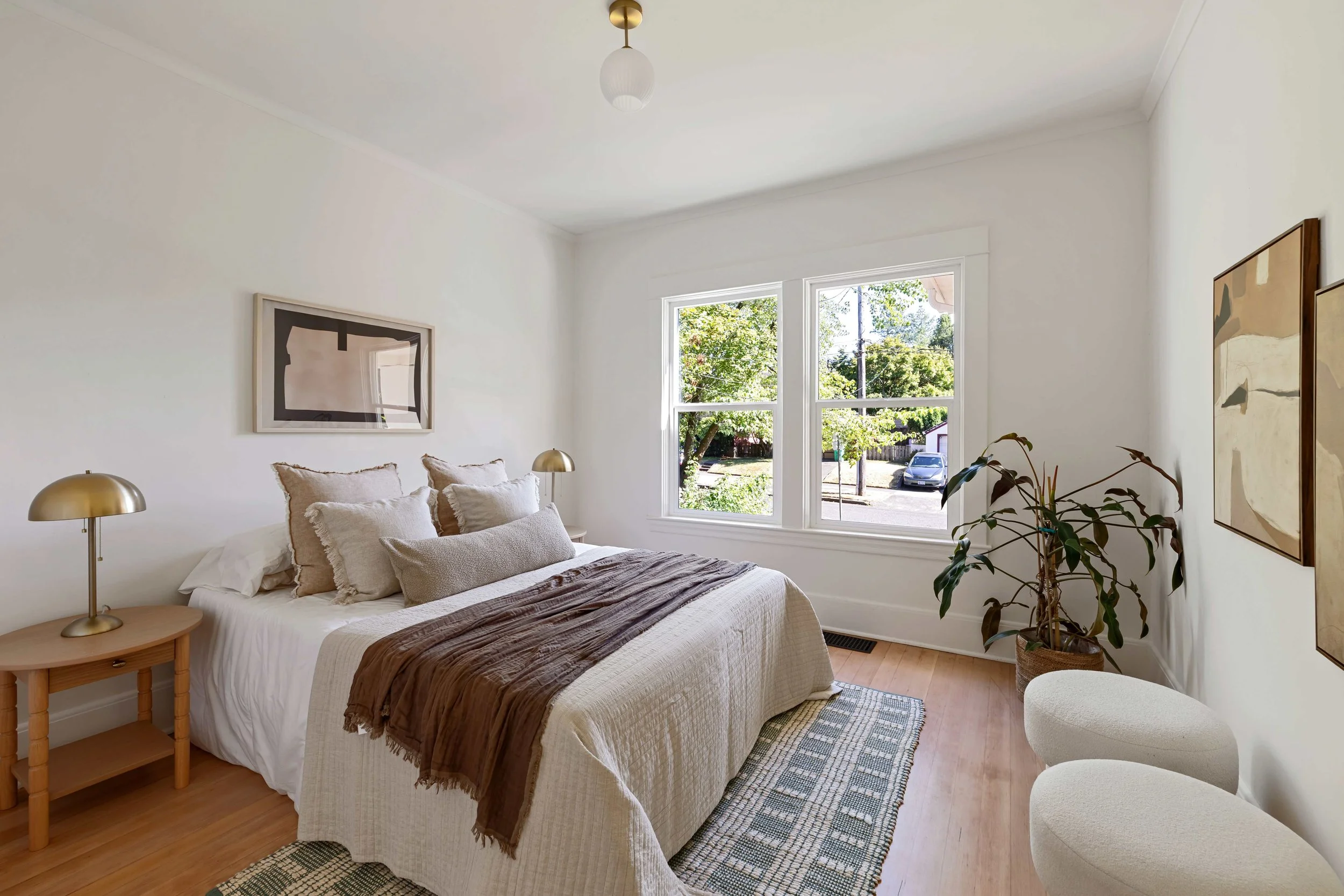 Bright bedroom with a large bed covered with white and beige bedding, a brown blanket, and multiple pillows. Two nightstands with gold lamps flank the bed. There are two framed artworks on the white walls and large windows showing greenery outside. The room has light hardwood floors and a patterned rug.