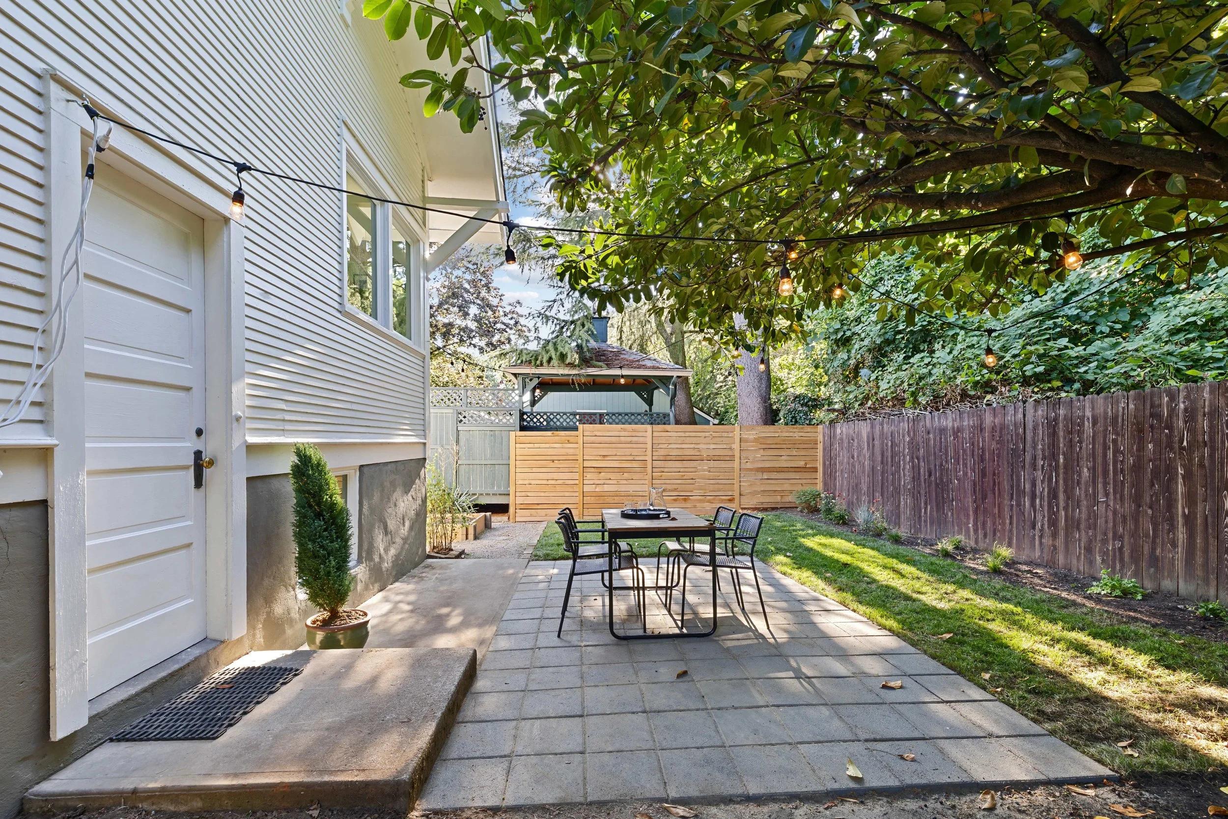 A backyard patio with outdoor dining set, surrounded by a lawn and wooden fences, shaded by a large leafy tree with string lights hanging across.