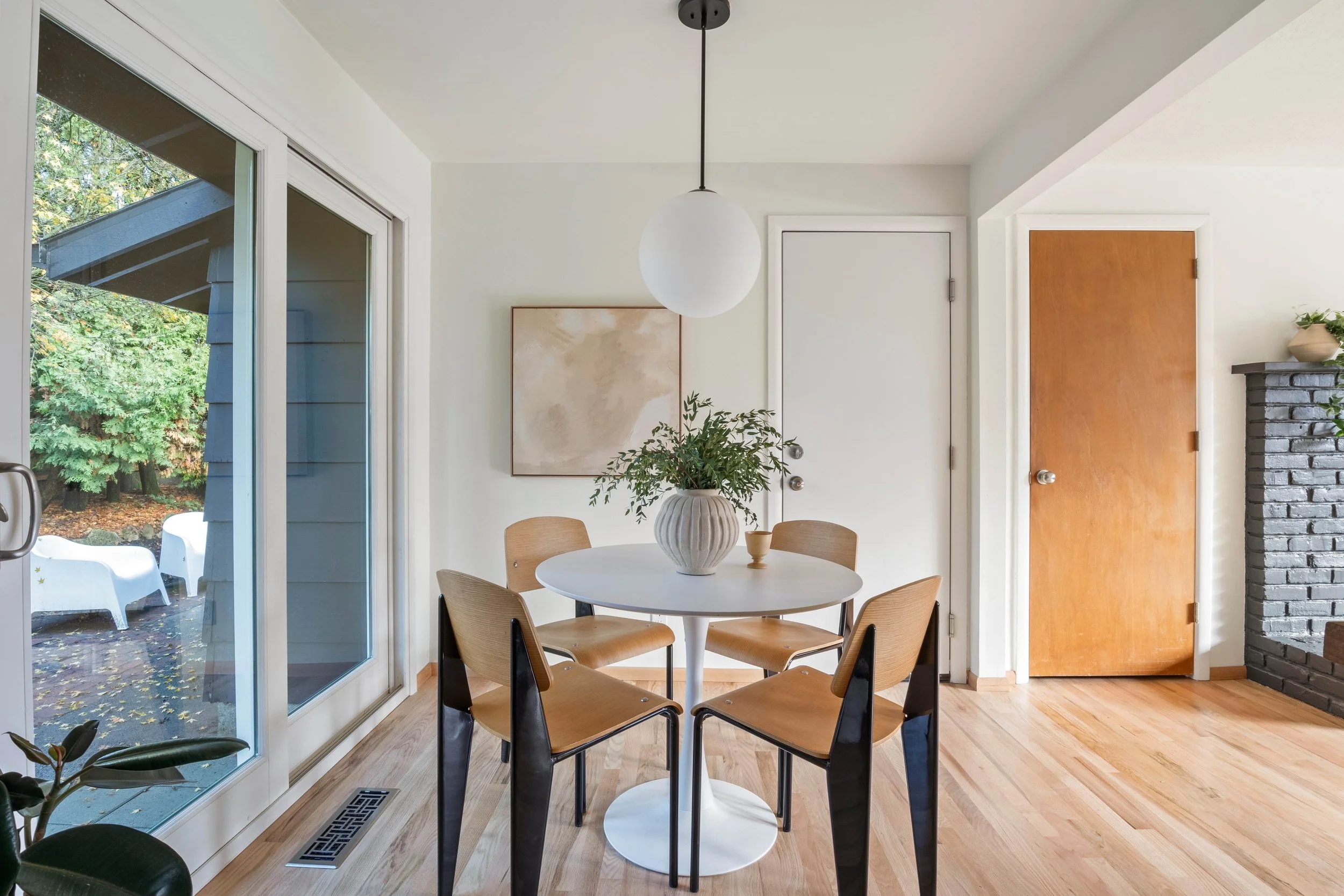 A cozy dining area with a round white table, four wooden chairs, a potted plant on the table, large sliding glass doors showing an outdoor garden with trees, a ceiling light fixture, a beige abstract artwork on the wall, a white door, and part of a brick fireplace.