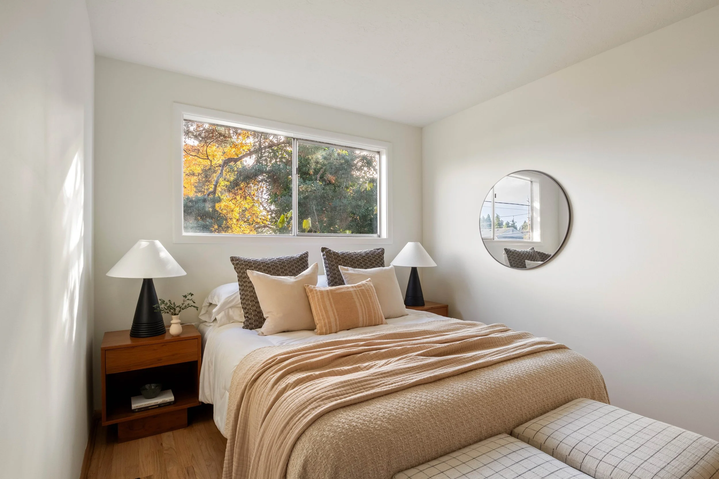 A cozy bedroom with a bed covered in beige and white bedding, decorative pillows, wooden nightstands with white table lamps, a small plant, a mirror on the wall, and a large window showing trees with autumn leaves.
