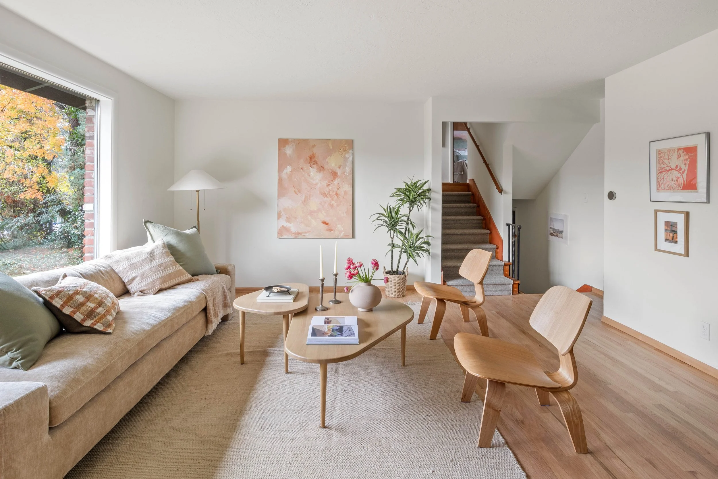 Living room with beige sofa, wooden coffee table, chairs, artwork on white walls, staircase, and large window showing trees with fall foliage.