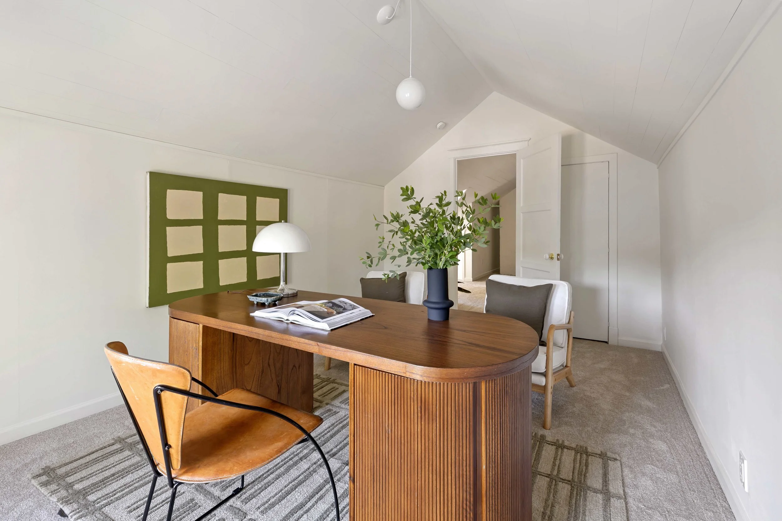 A dining room with a wooden table, white chairs, a green abstract art piece, a white lamp, and a potted plant, with white walls and a sloped ceiling.