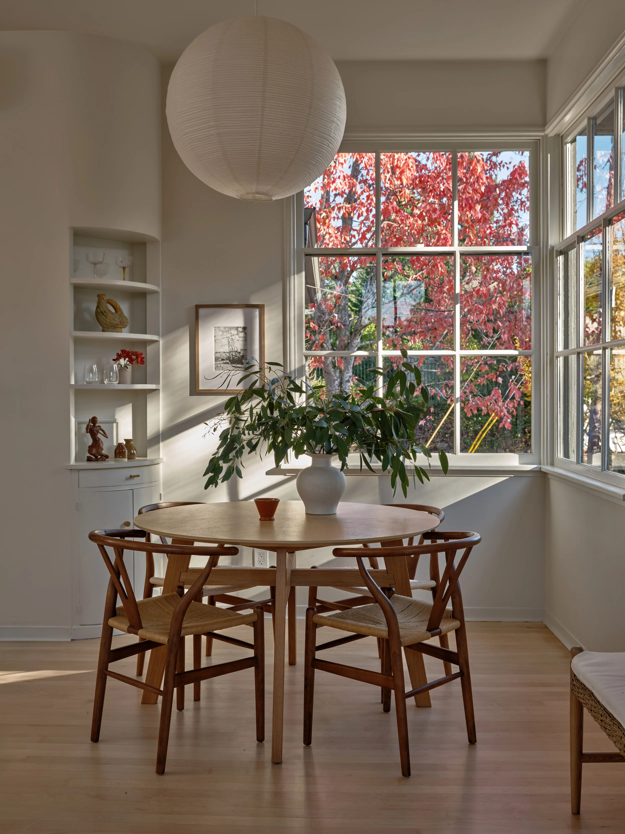 Dining room with a round wooden table, six wooden chairs, a large white ceramic vase with green plants, large windows showing red autumn leaves, a white wall with built-in shelves, and a paper lantern ceiling light.