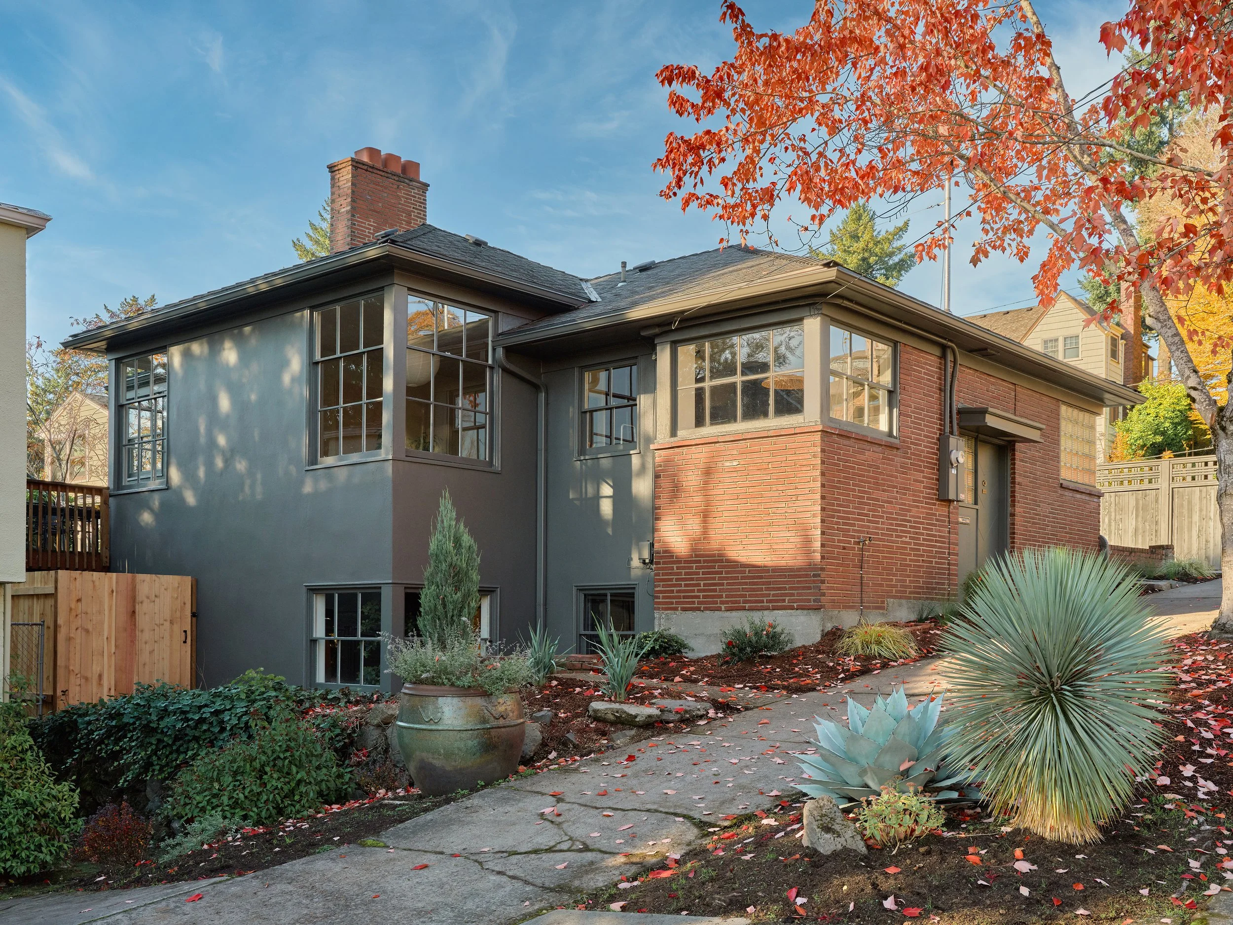 A house with large windows, a brick chimney, and a garden with plants and a pathway, under fall foliage with orange leaves.