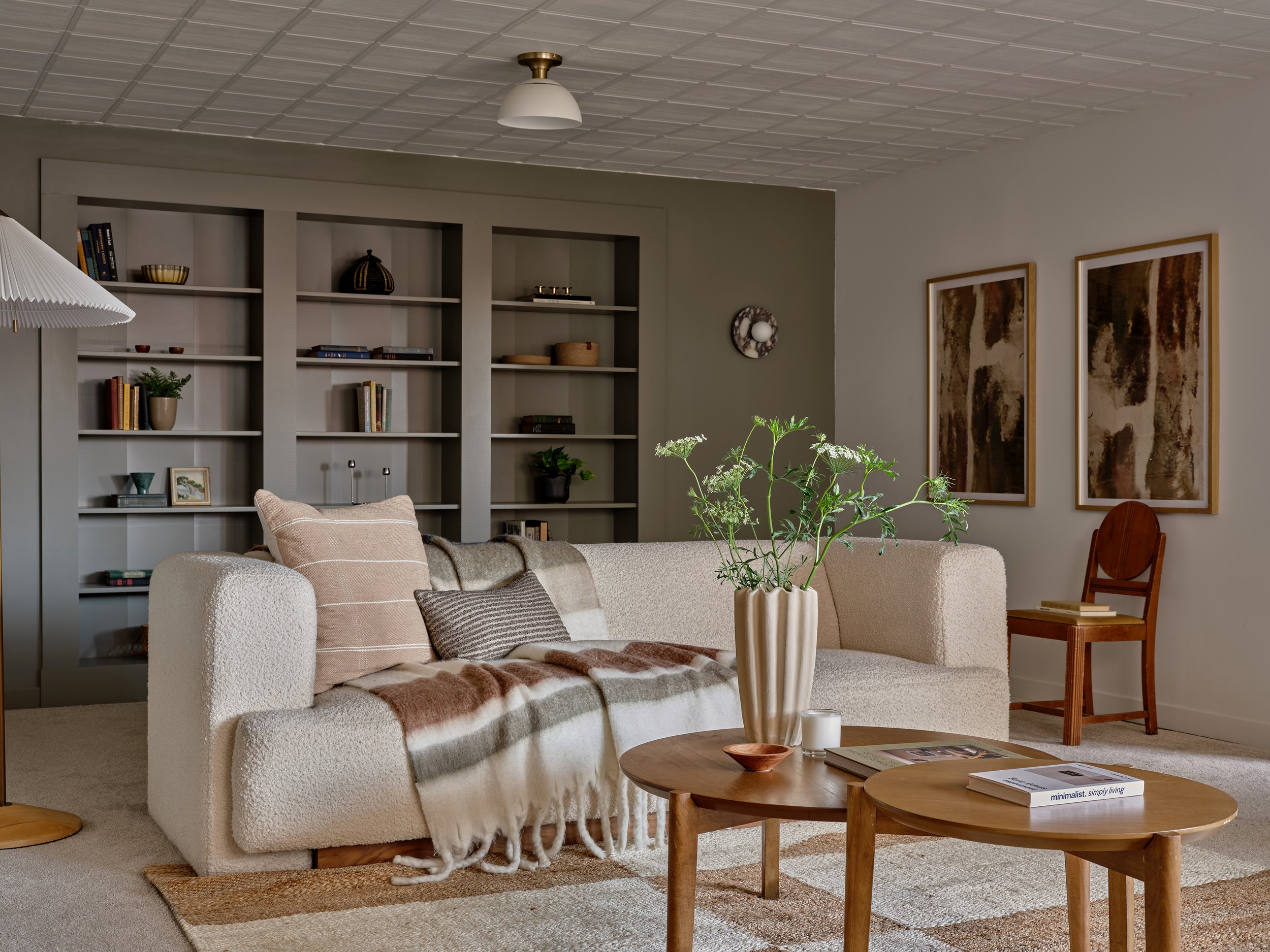A cozy living room with a white textured sofa, beige and brown striped and throw pillows, a blanket, and a coffee table with books, a small bowl, and candles. There is a plant in a white vase on the coffee table. Behind the sofa, there's a built-in bookshelf filled with books, decor, and a potted plant. To the right of the bookshelf, there are two abstract art pieces framed on the wall. The ceiling is tiled, and a small ceiling light is visible. The room has a warm, inviting ambiance with earthy tones and natural decor.