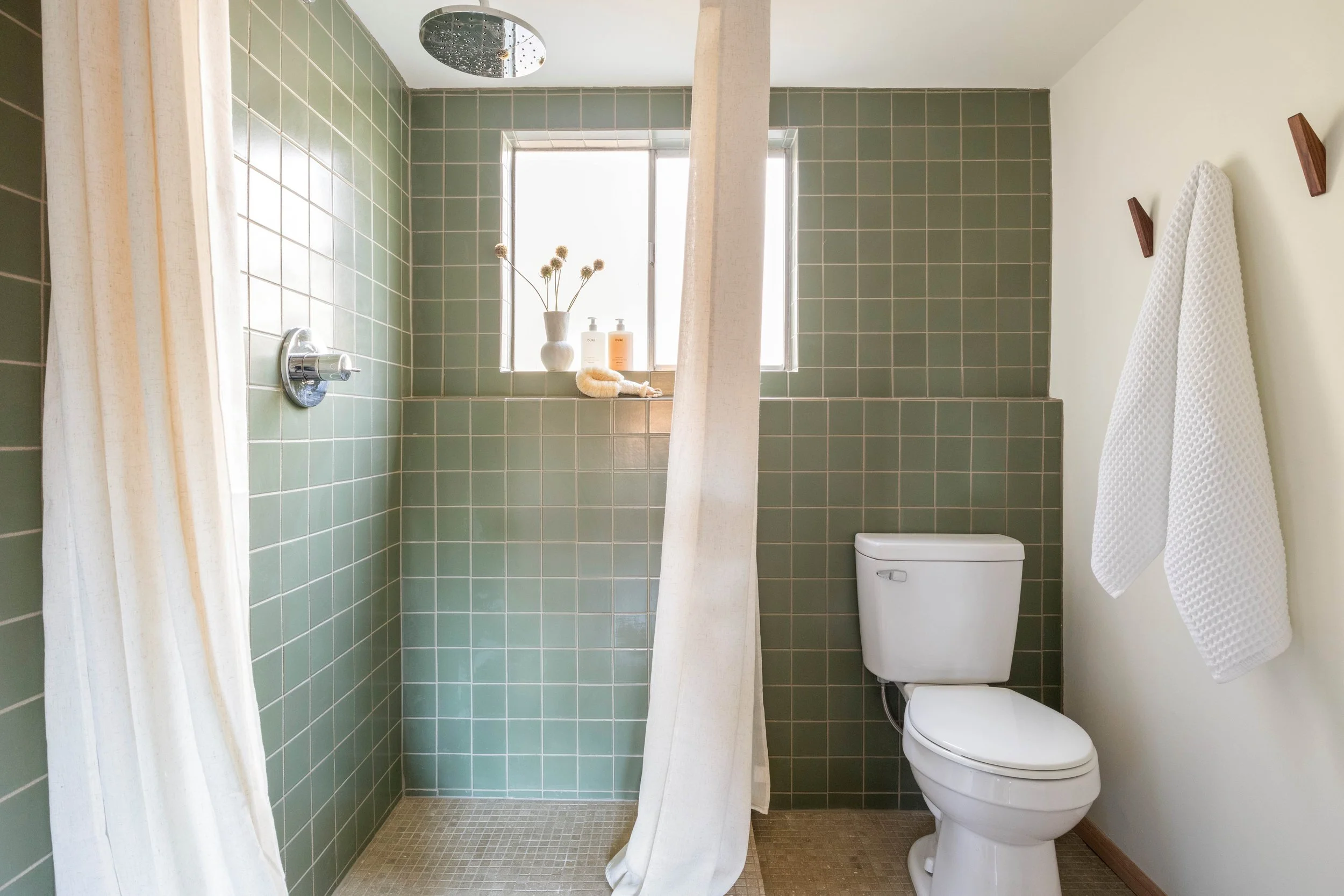 A bathroom with green tiled walls, a shower with a rain shower head, a white curtain, a toilet, a shelf with a vase and toiletries, a window, and a white towel hanging on a wooden hook.