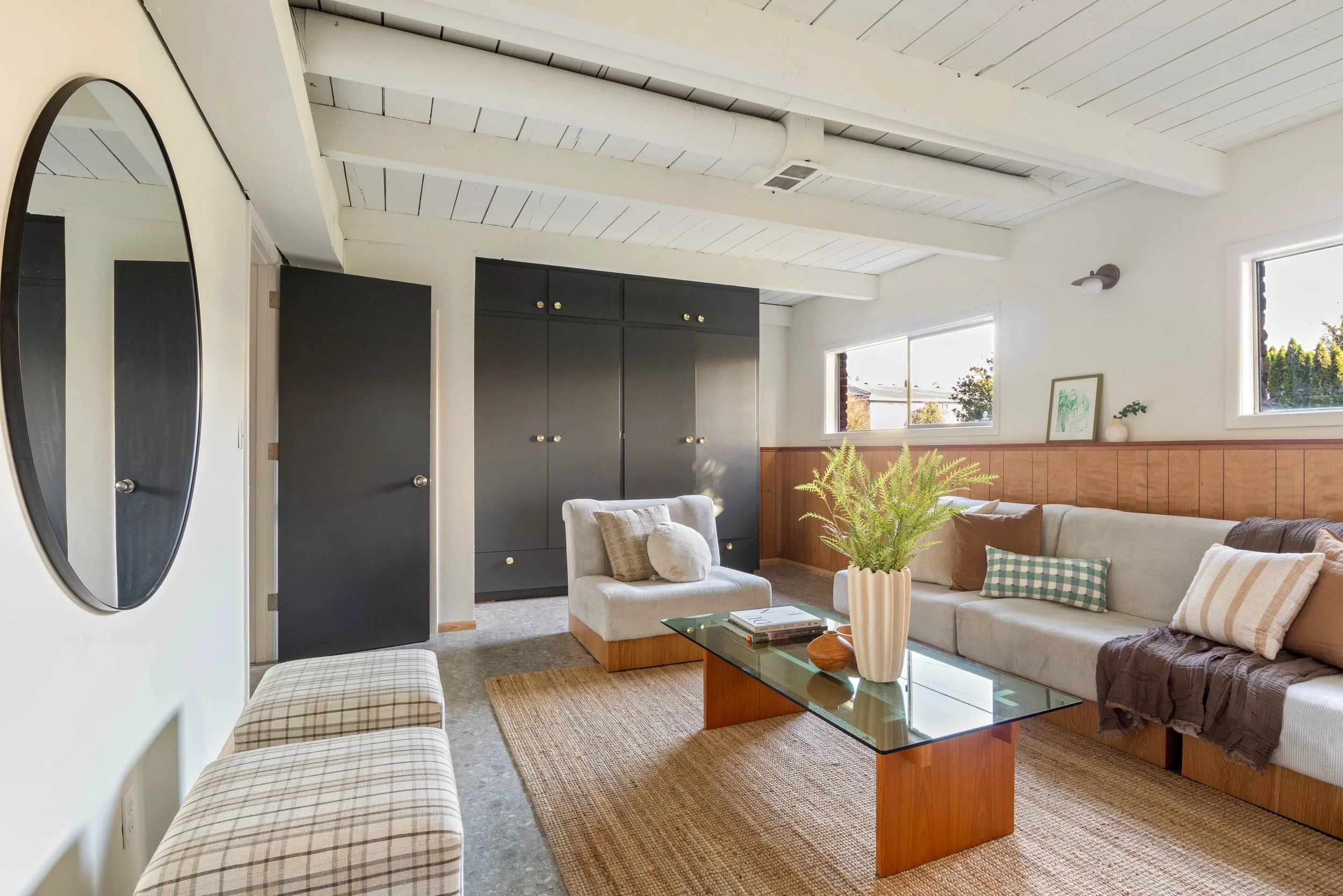 Living room with beige sofa, plaid armchair, glass coffee table, black storage cabinet, oval mirror, potted plant, framed art, large windows, white ceiling with exposed beams.