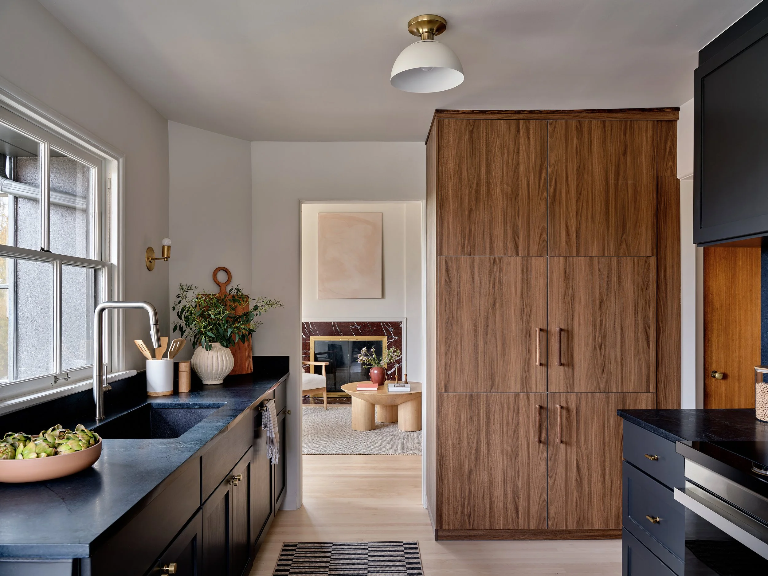 A modern kitchen with black cabinets, a large black countertop, a window with white framing, and a wooden cabinet. The kitchen opens into a living room with a fireplace and framed artwork. The room has light wood flooring, and there are decorative plants and accessories.
