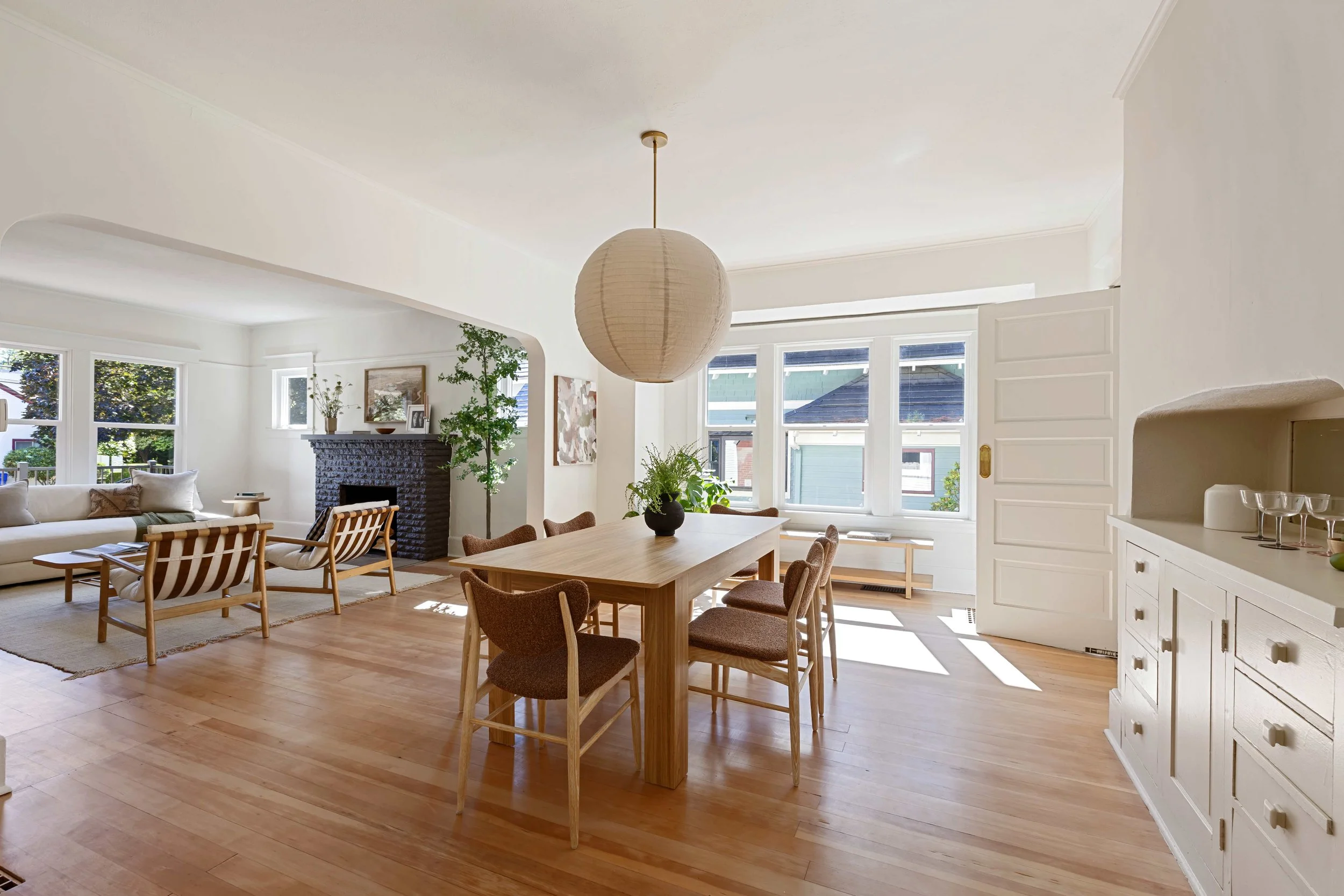 A spacious, bright living room and dining area with large windows, wooden flooring, a white sofa, wooden chairs, a black brick fireplace, and various decorative items including plants, paintings, and a paper lantern pendant light.