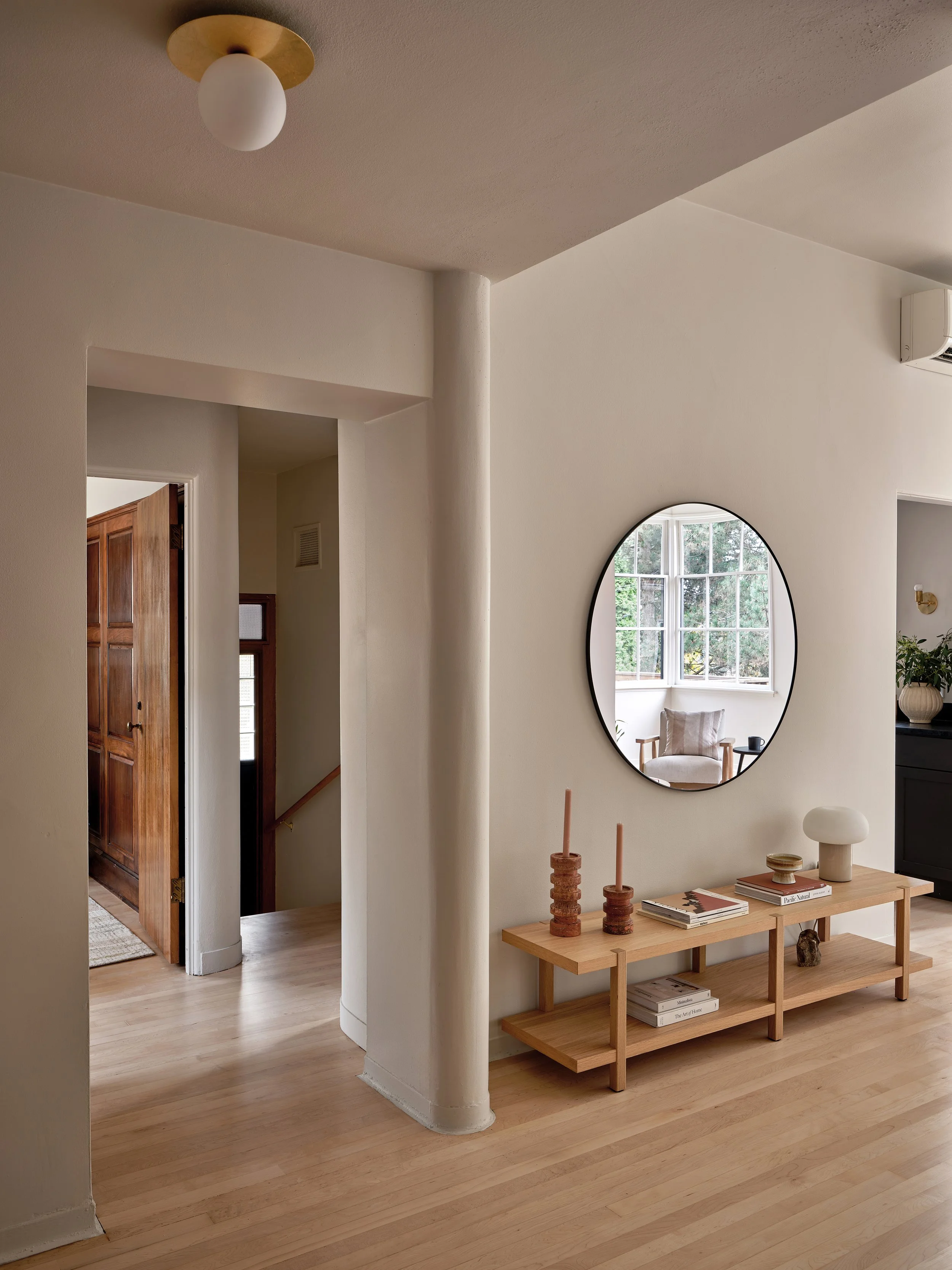 Interior home entryway with a door, a rounded mirror on the wall reflecting a window and a chair, and a wooden console table decorated with two candles, books, and small decorative objects.