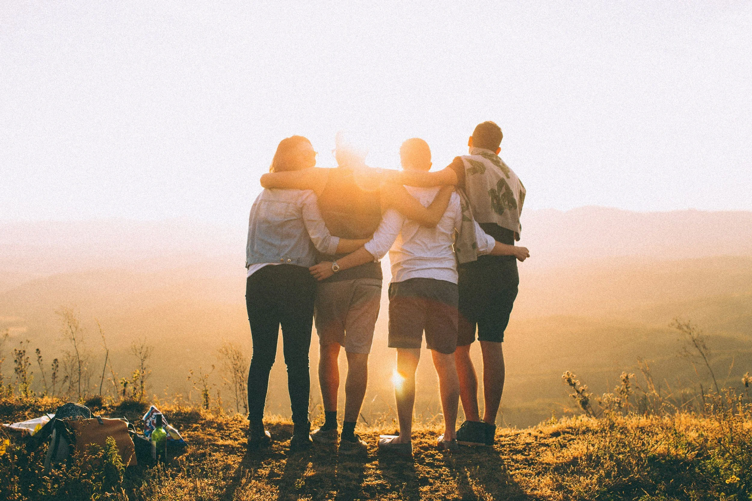 Group of five young adults standing in a row with arms around each other, watching a sunset in an outdoor field.