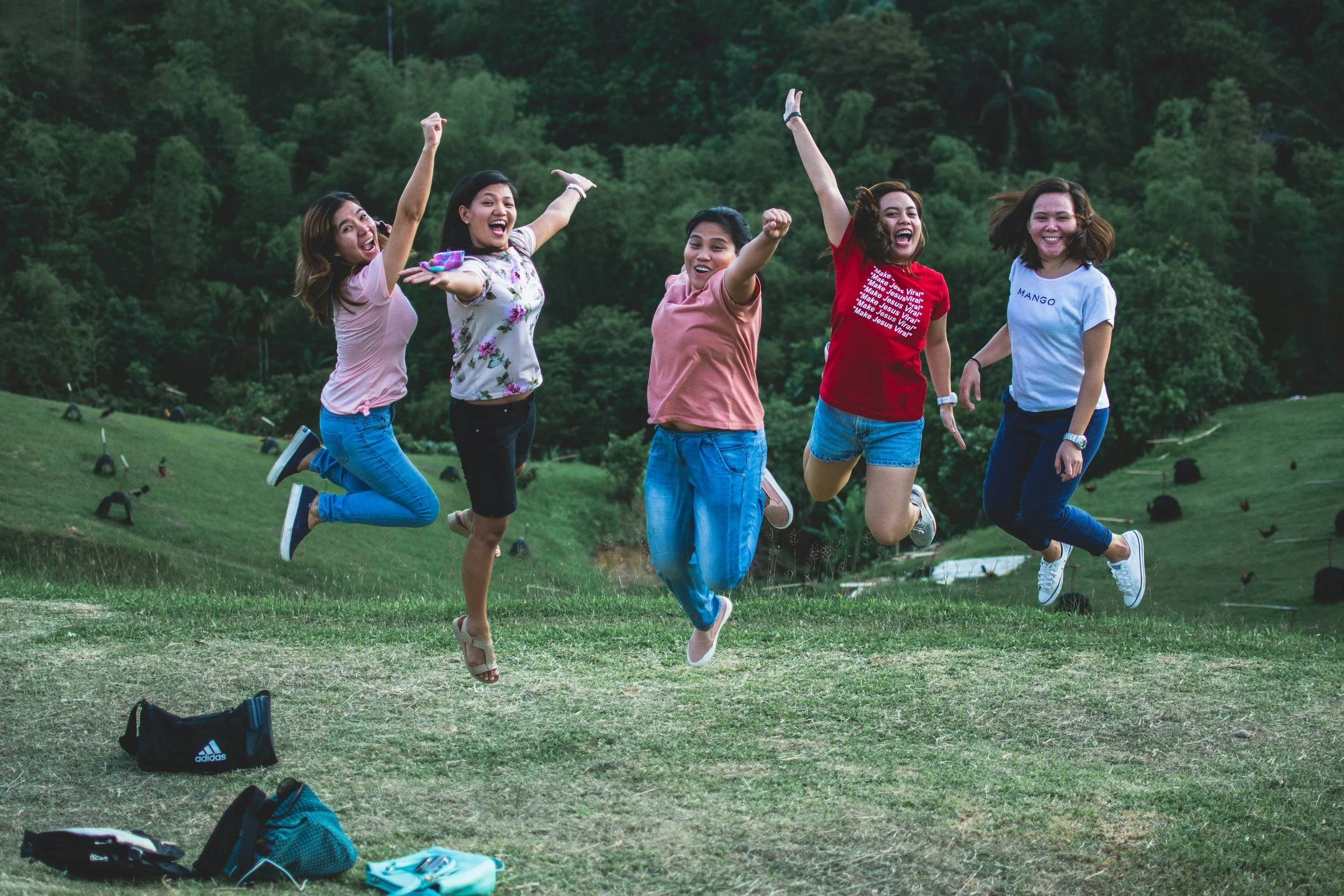 Group of five young adults jumping and smiling in a grassy outdoor area with trees in the background.
