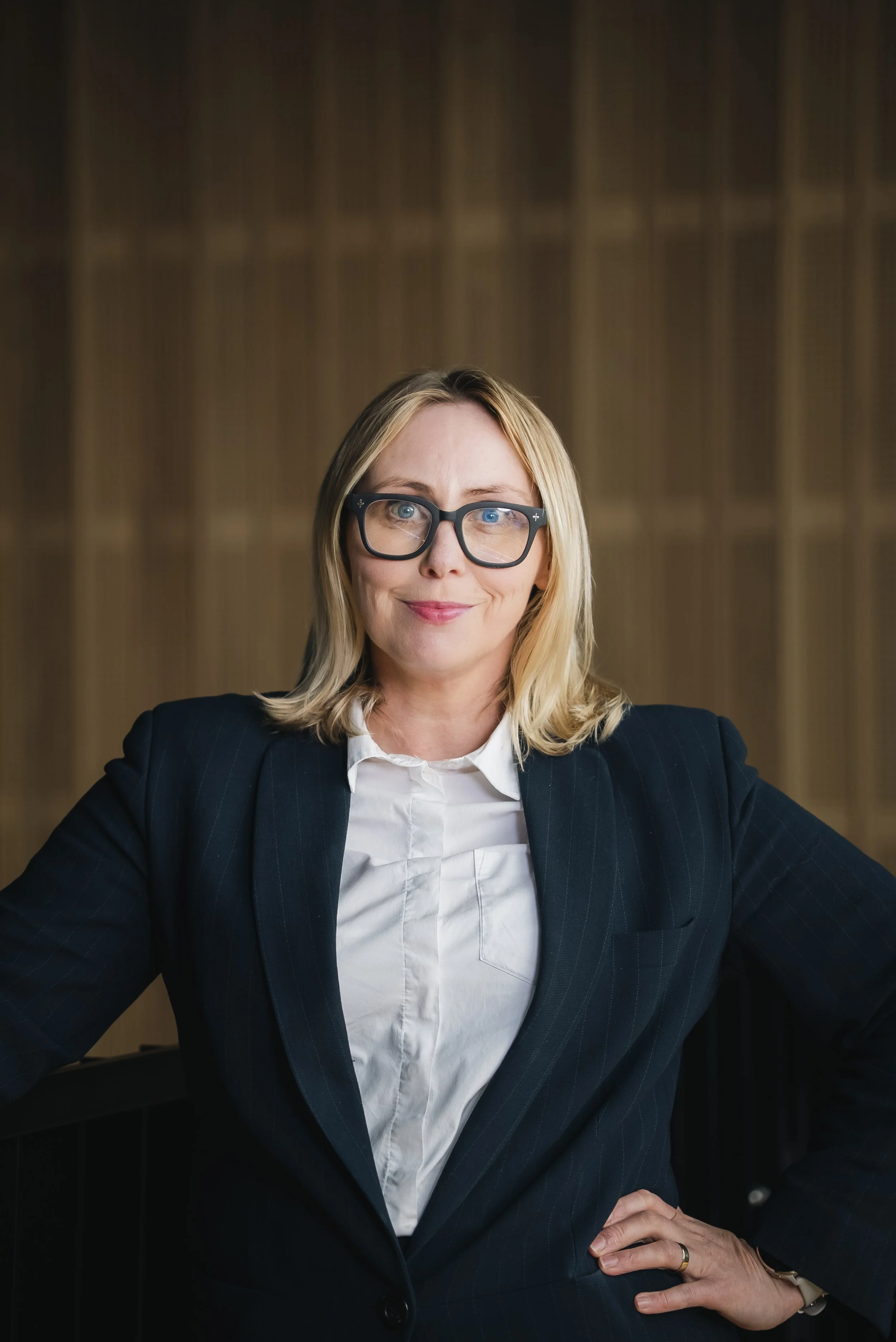 A woman with blonde hair, wearing glasses, a dark pinstripe suit, and a white shirt, sitting confidently at a desk with her hands on her hips, against a dark wooden-paneled background.
