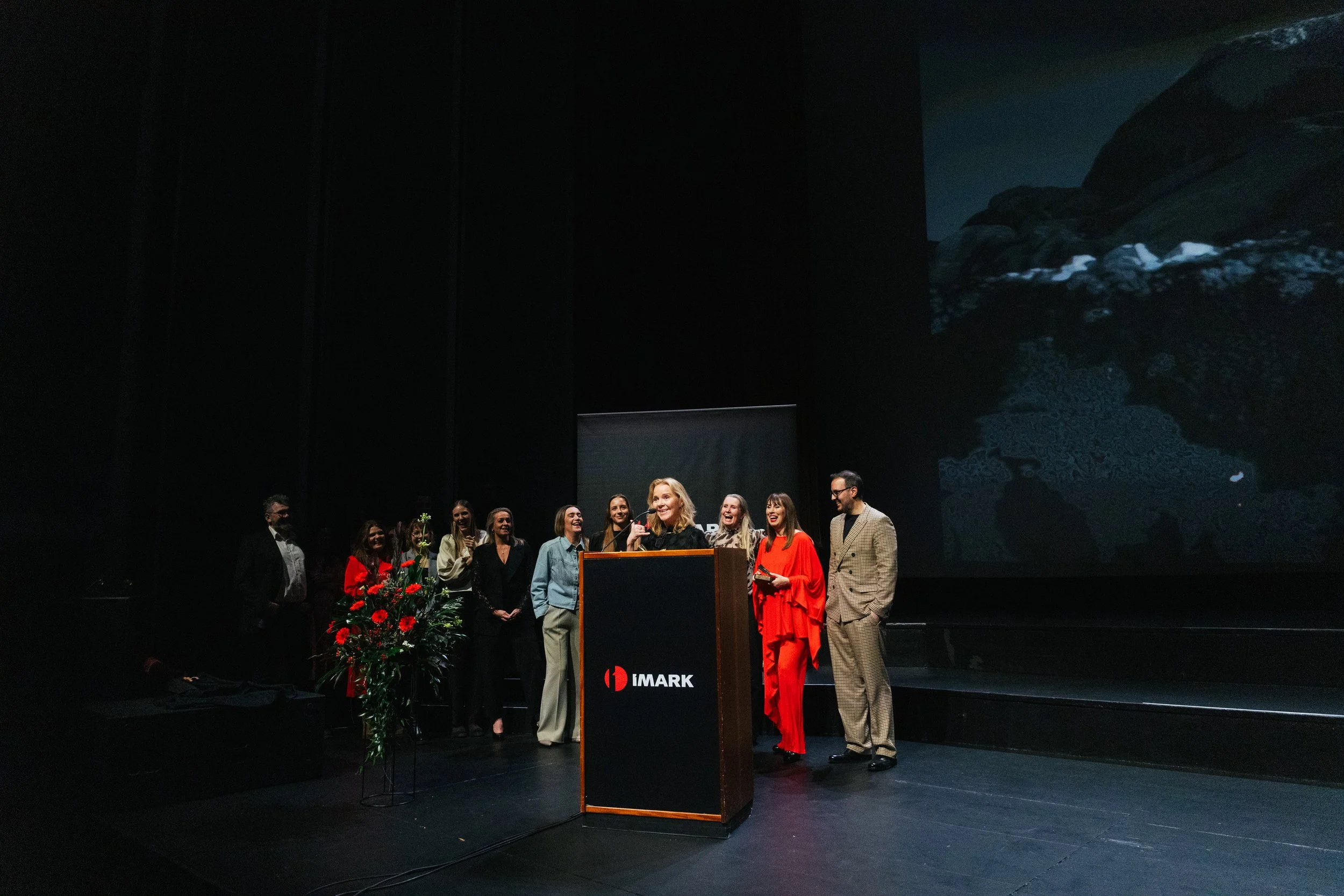 A group of people on stage at an awards ceremony, with a woman speaking at a podium that has the IMARK logo, and a large screen behind showing a mountain landscape.