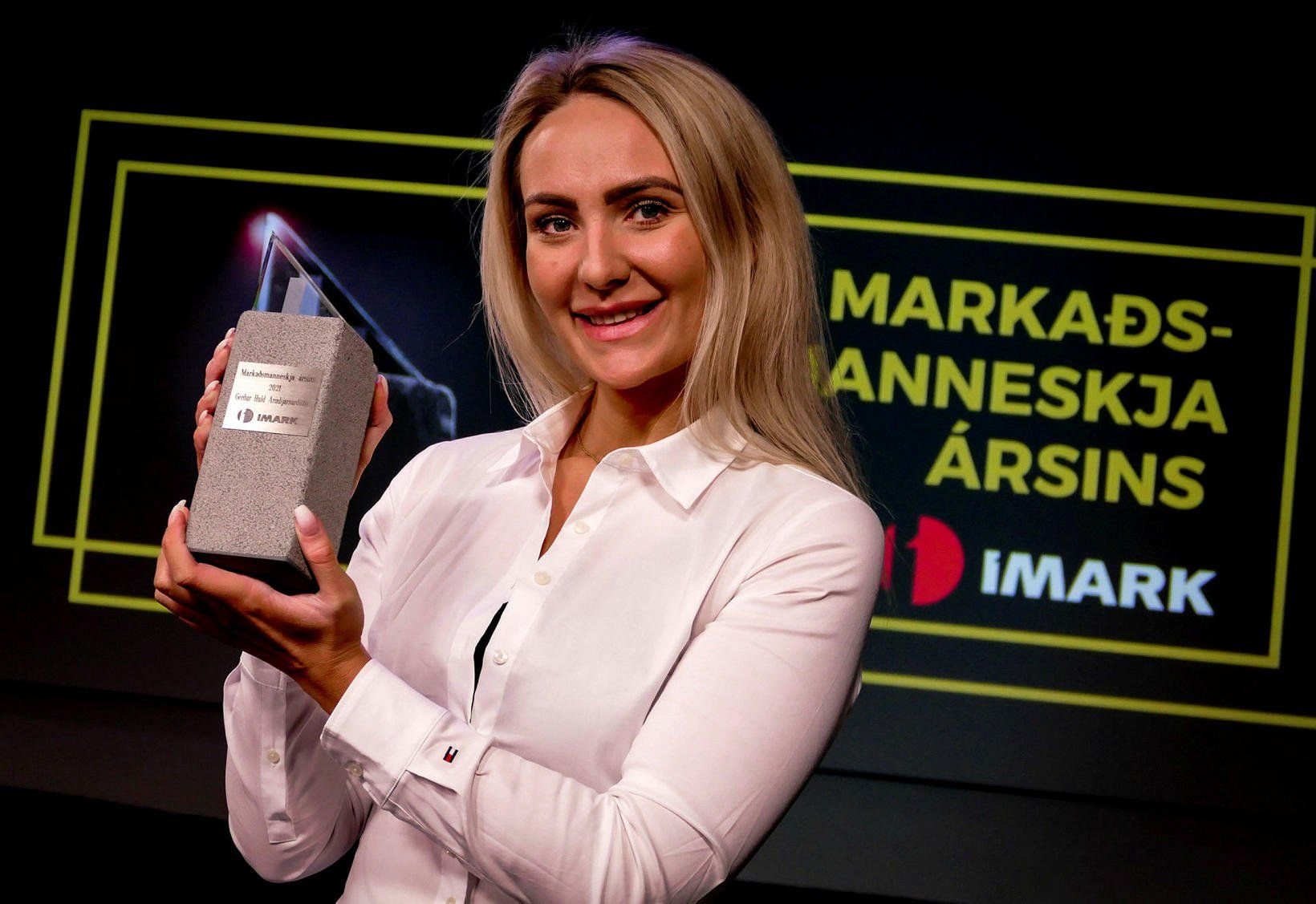 A woman with long blonde hair holding an award in front of a black background with yellow text and a logo, smiling at the camera.