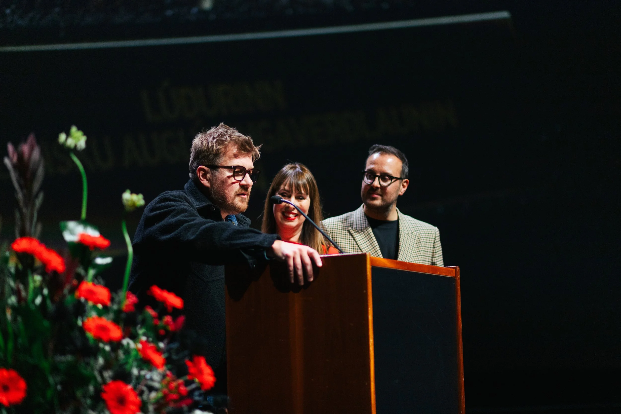 Three people, two men and a woman, standing behind a podium with flowers in the foreground. The man on the left has glasses, curly hair, a beard, and is wearing a black coat. The woman in the middle has long hair and is smiling. The man on the right 