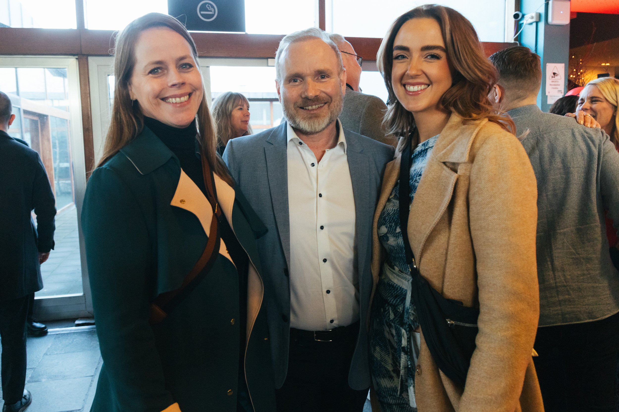 Three people smiling and posing for a photo indoors. Two women on either side of a man in a suit. Multiple people are in the background, and there are large windows with daylight.