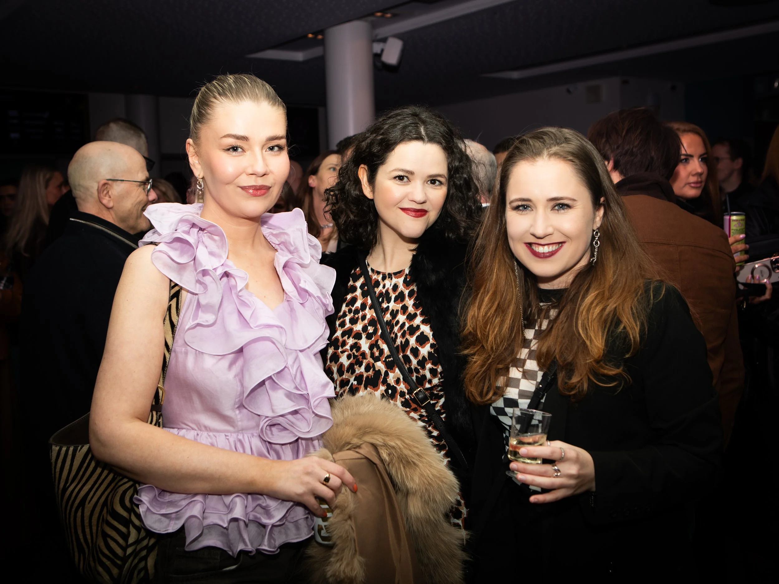 Three women standing together at a social event, smiling, with the woman on the right holding a glass of champagne. They are dressed up and surrounded by other attendees in a dimly lit indoor venue.
