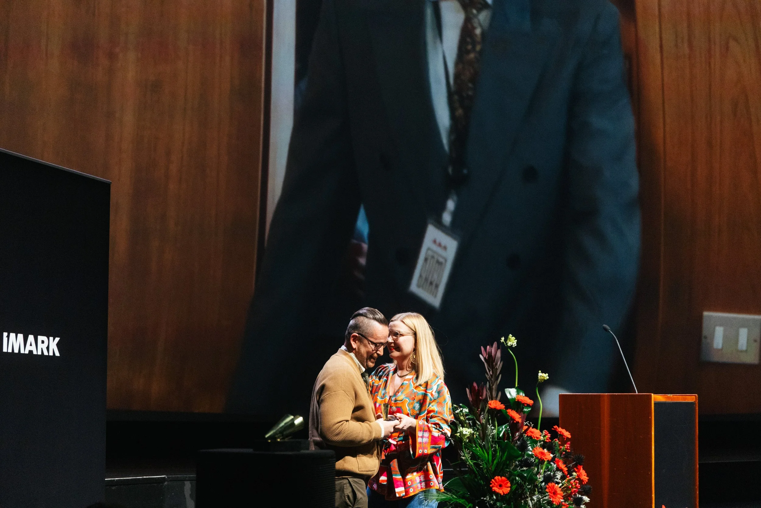 At an event, a woman and a man are exchanging a handshake on stage with flower arrangements nearby, while a large projection screen shows a close-up of a couple in the background.
