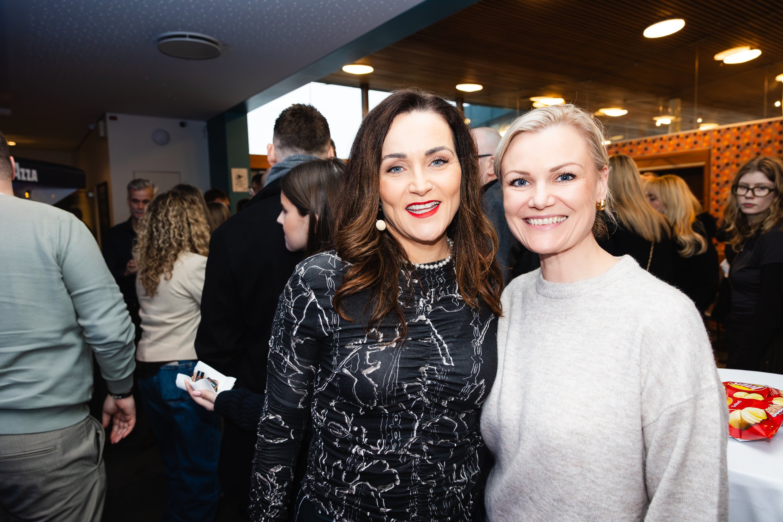 Two women are smiling and posing for the camera at a crowded indoor event. One woman has dark hair and wears a patterned black and white top, and the other has blonde hair and wears a light gray sweater.