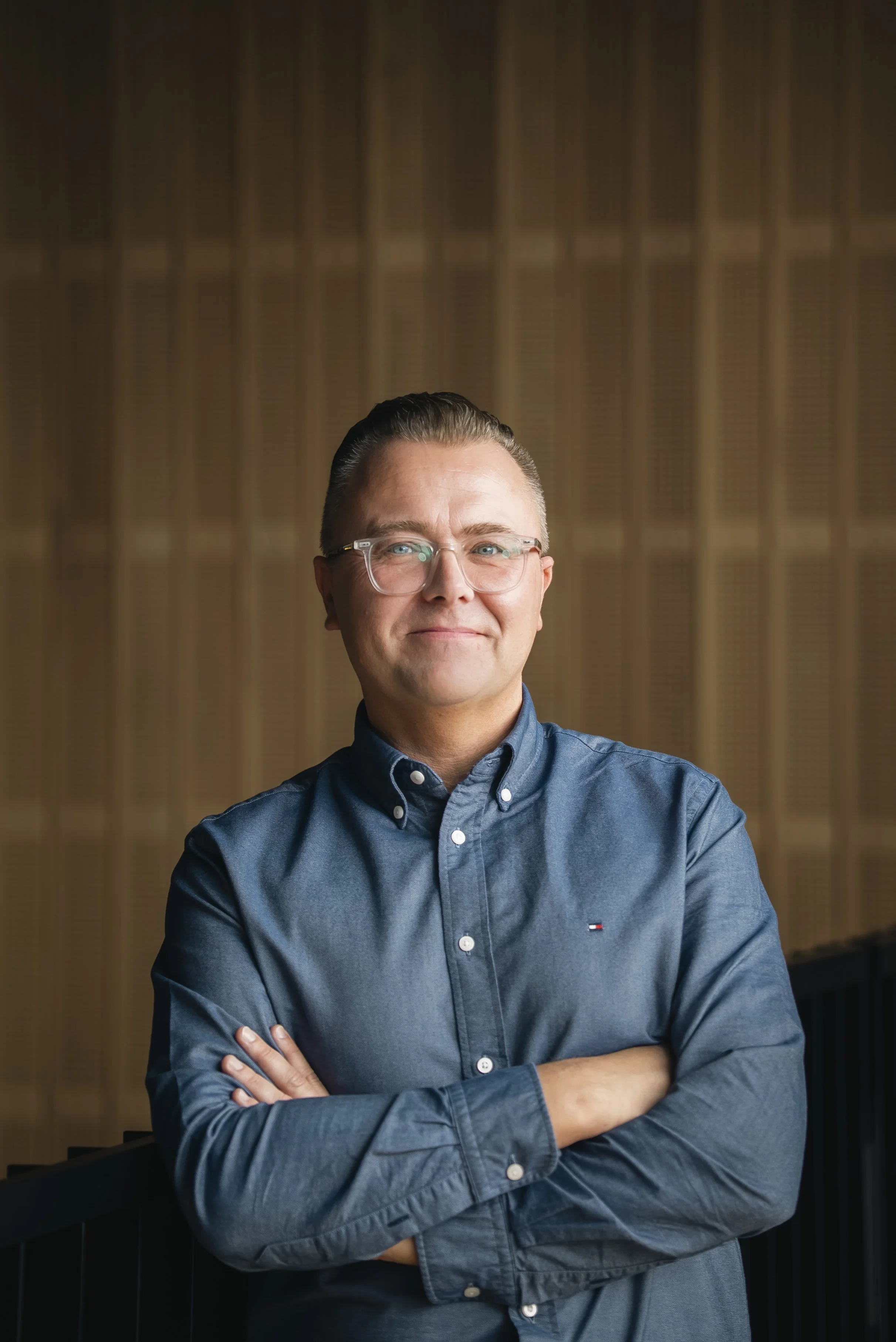 A man wearing glasses and a blue button-up shirt, smiling with arms crossed, standing in front of a wooden background.
