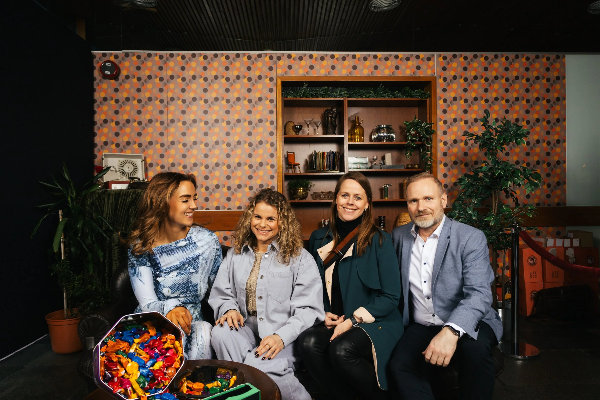 Group of four people seated together indoors, smiling at the camera. There are plants and a decorated wall with shelves in the background, and a container of colorful wrapped candies on the table in front of them.