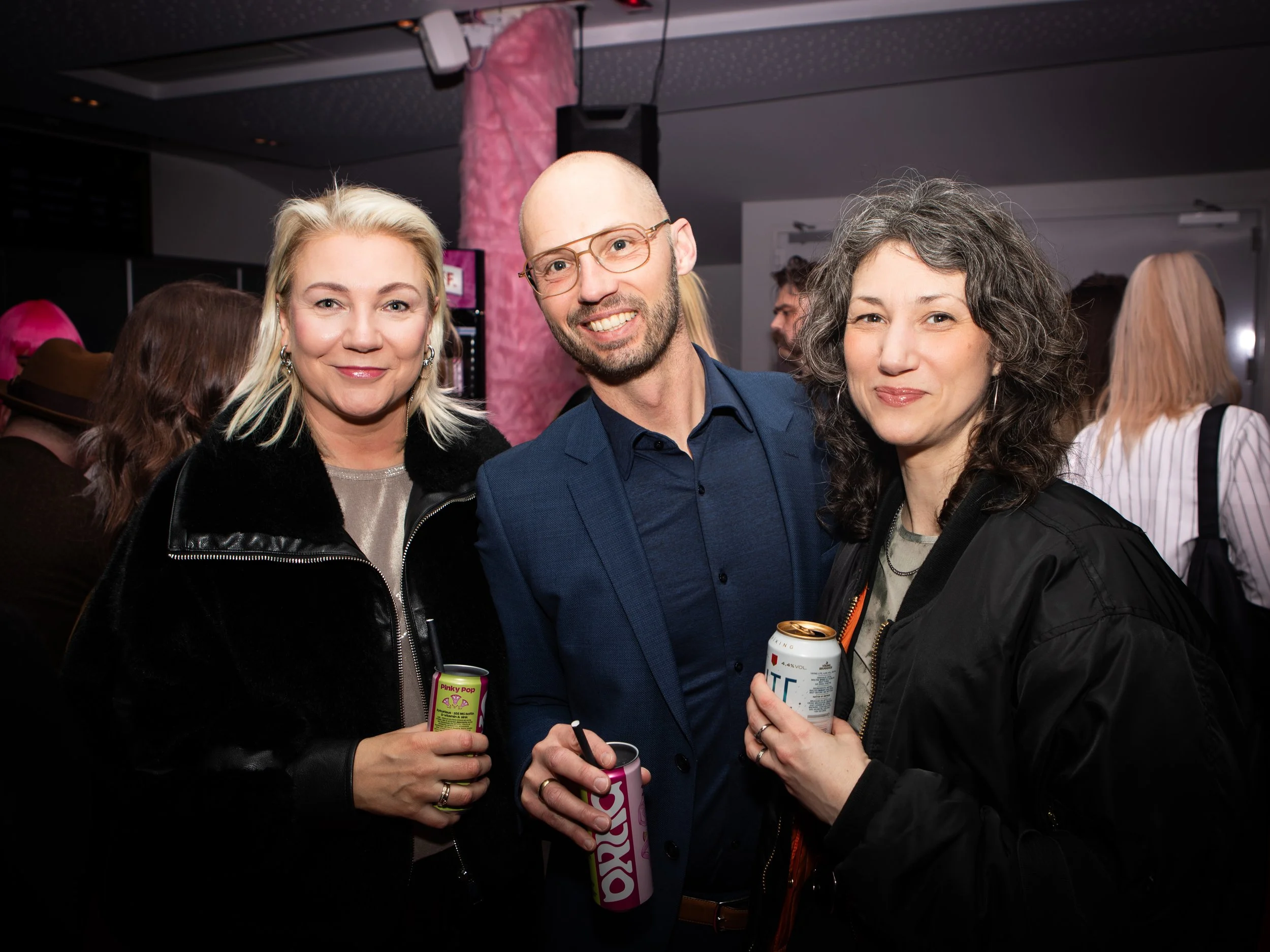 Three people are smiling and holding drinks at a social gathering. They appear to be at a lively indoor event with other attendees in the background.